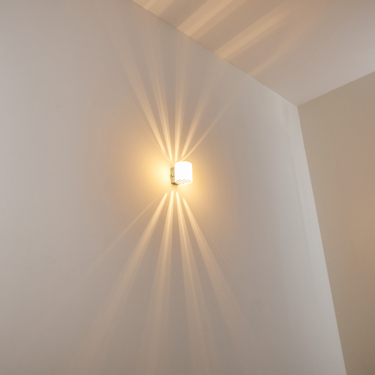 Eine Wandleuchte wirft Lichtstrahlen auf eine weiße Wand. Das Licht ist rechteckig mit gewölbter Oberseite. Die Wand ist hell.