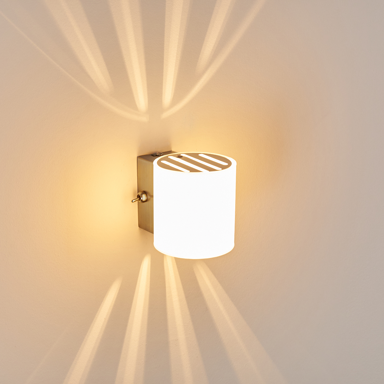 Eine zylindrische Wandleuchte mit Lichtstrahlen an einer beigen Wand. Die Lampe ist weiß mit einem Metallfuß.