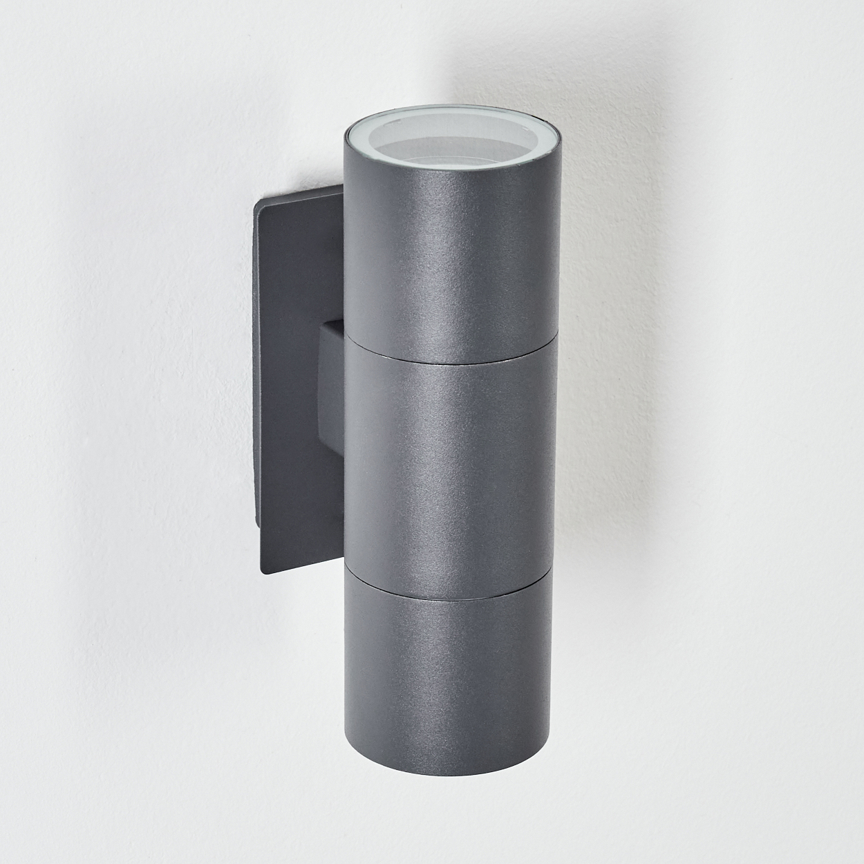 Eine graue, zylindrische Wandleuchte ist an einer weißen Wand montiert. Die Lampe hat eine klare Oberseite und einen rechteckigen Sockel.