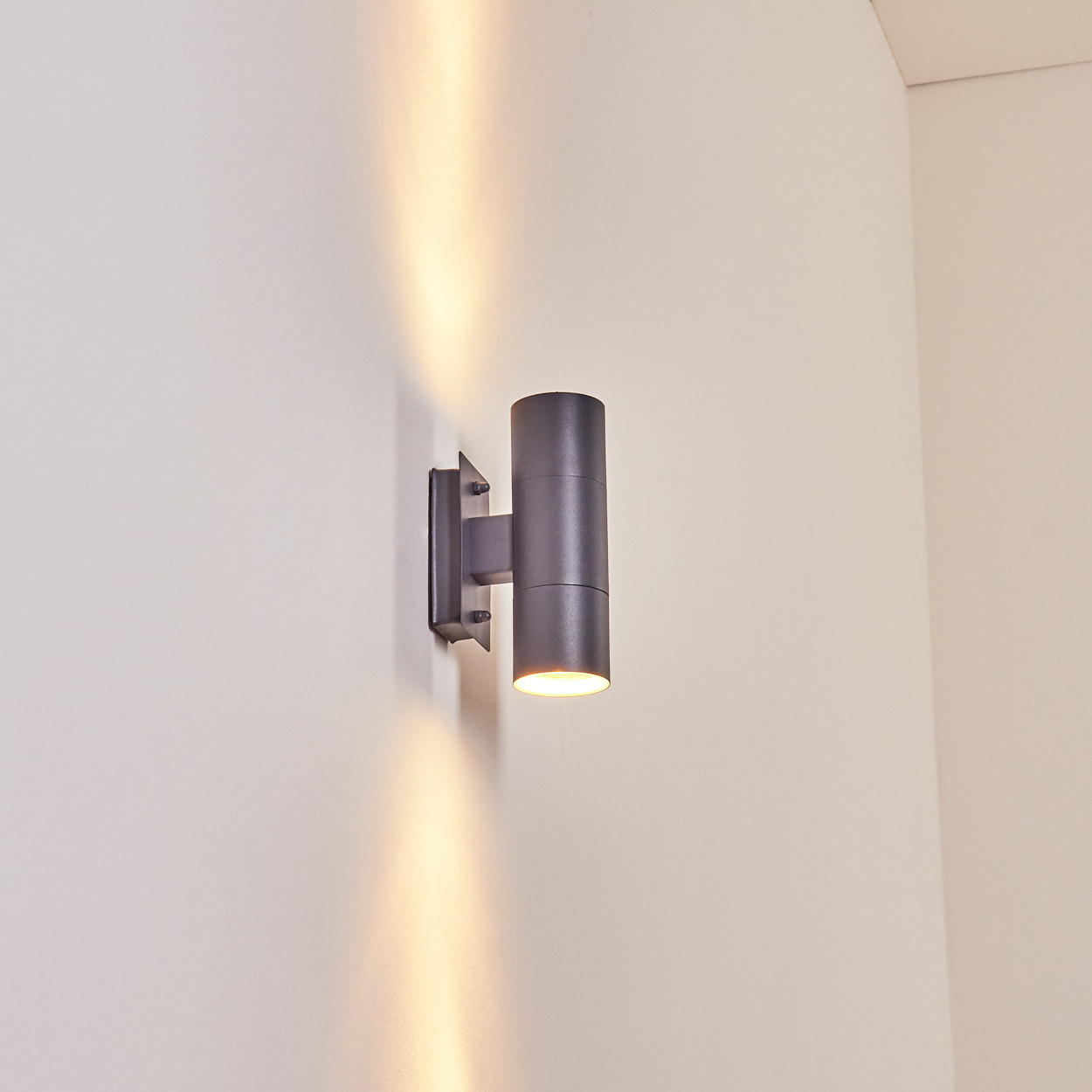 Dunkelgraue, zylindrische Außenwandleuchte an hellfarbener Wand. Lichtstrahlen gehen davon aus.