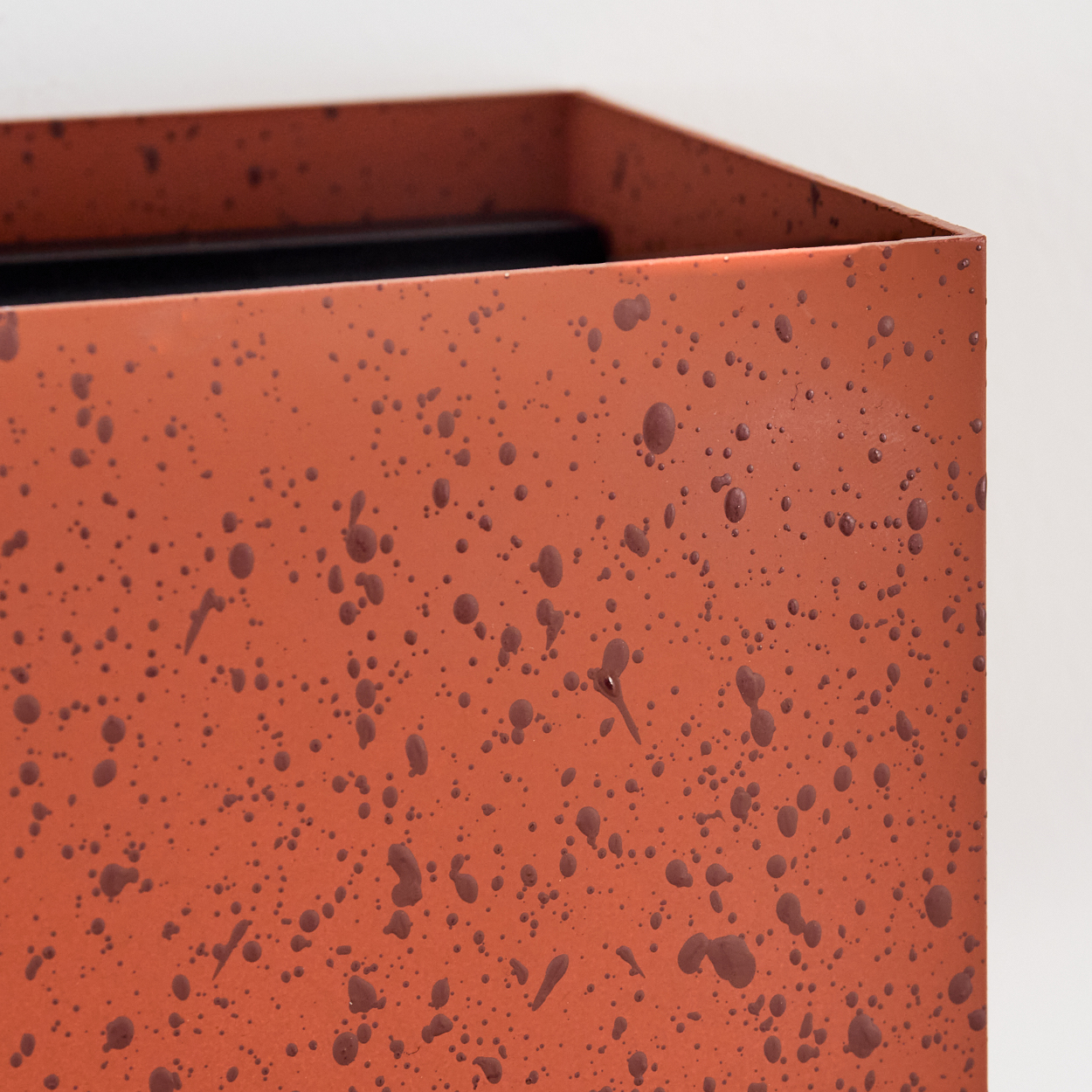 Nahaufnahme einer rostorangefarbenen Metallbox mit dunkelbraunen Farbspritzern. Der Hintergrund ist schlicht weiß.