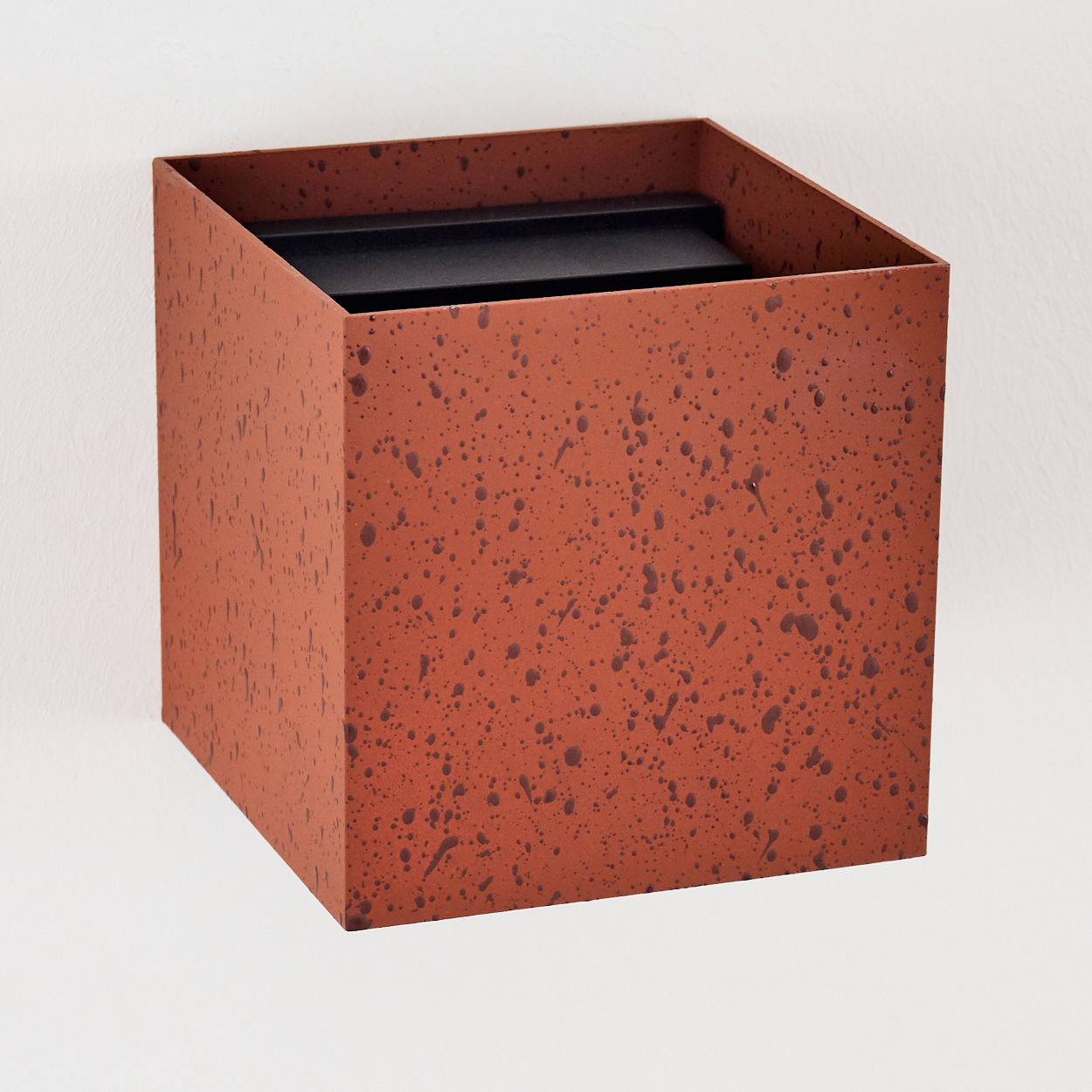 Ein quadratischer Behälter mit offener Oberseite. Er ist rostfarben mit dunkleren Sprenkeln. Im Inneren des Behälters ist eine schwarze Oberfläche zu sehen.