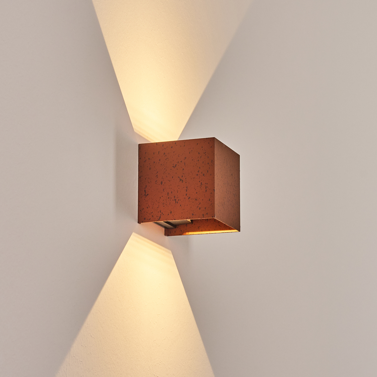 Eine quadratische, rostfarbene Wandleuchte strahlt Licht nach oben und unten an eine hellfarbene Wand. Die Lampe hat eine kastenförmige Gestalt.