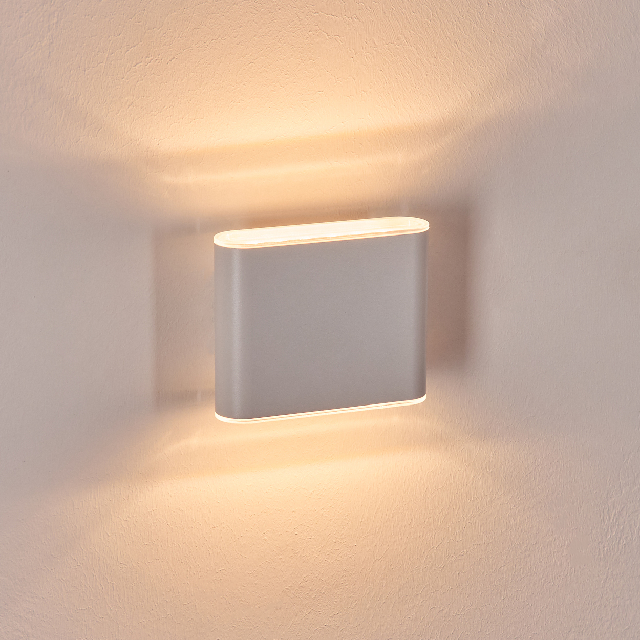 Eine moderne, graue Wandleuchte ist an einer strukturierten, cremefarbenen Wand montiert. Die Lampe strahlt warmes Licht aus und wirft Schatten.
