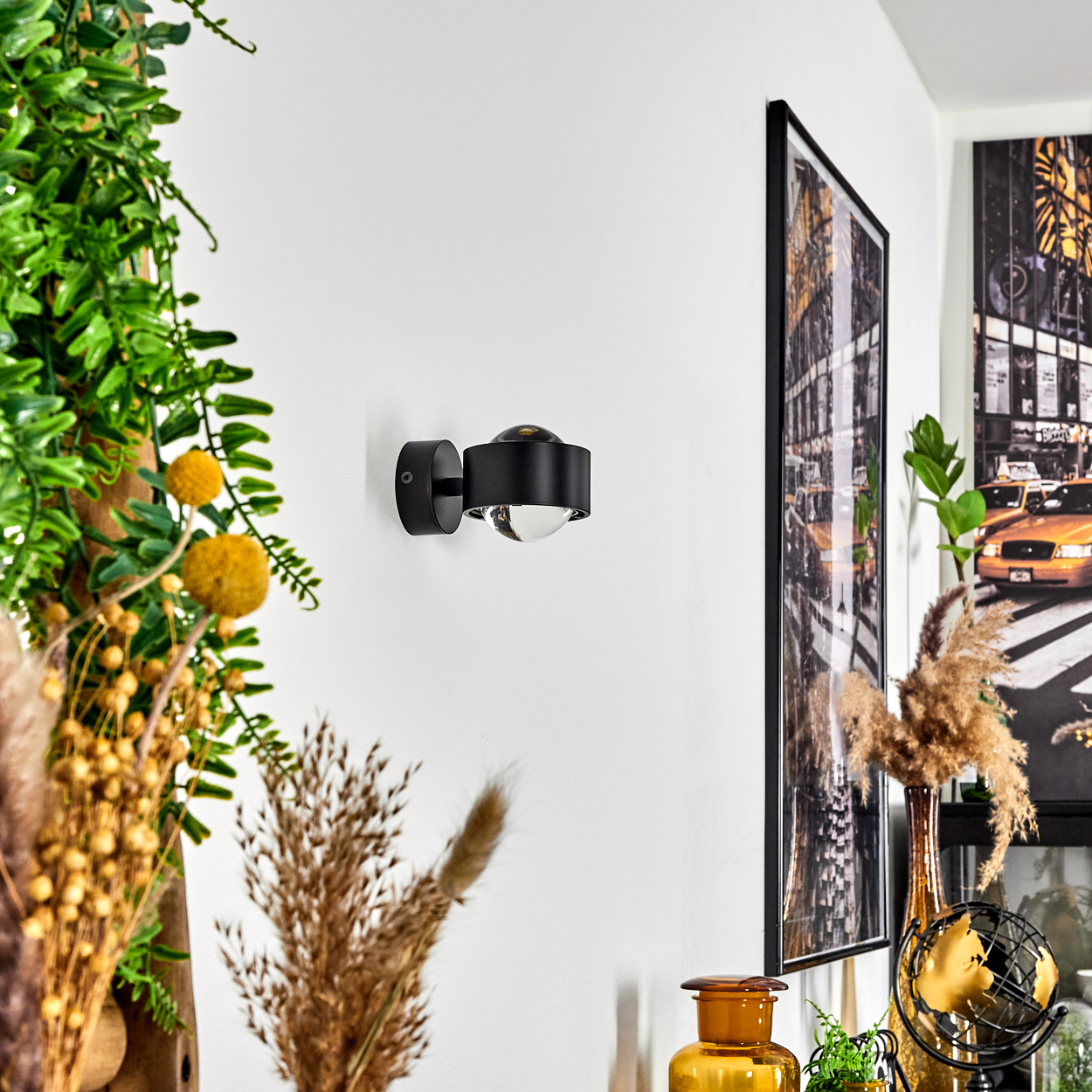 Eine moderne schwarze Wandleuchte mit klarer Glühbirne an einer weißen Wand. Weitere Dekorationen sind Pflanzen und gerahmte Kunstwerke.