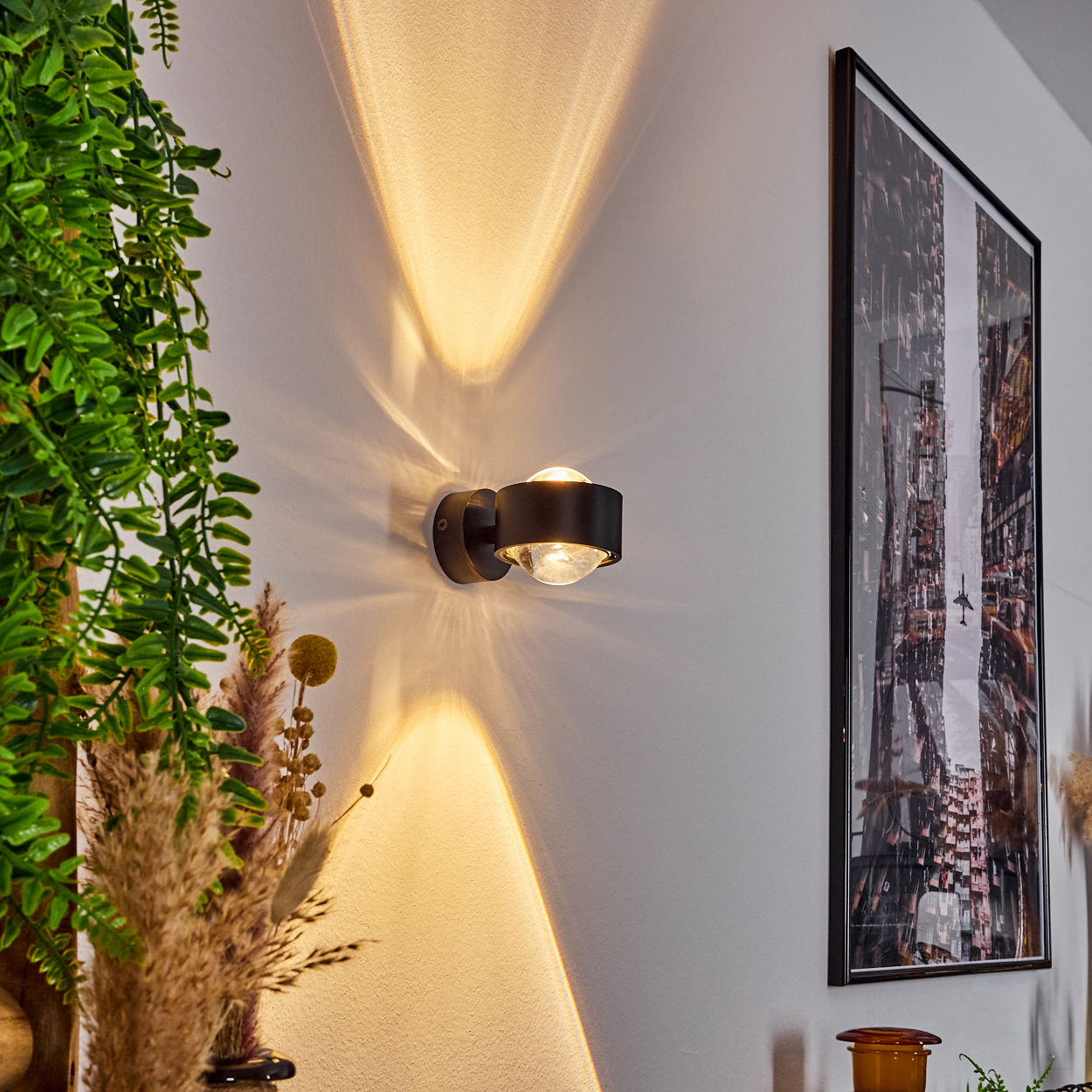 Eine moderne Wandleuchte strahlt Licht aus. Ein Bild einer Stadt hängt an der Wand. Grüne Pflanzen und Trockenblumen sind zu sehen.