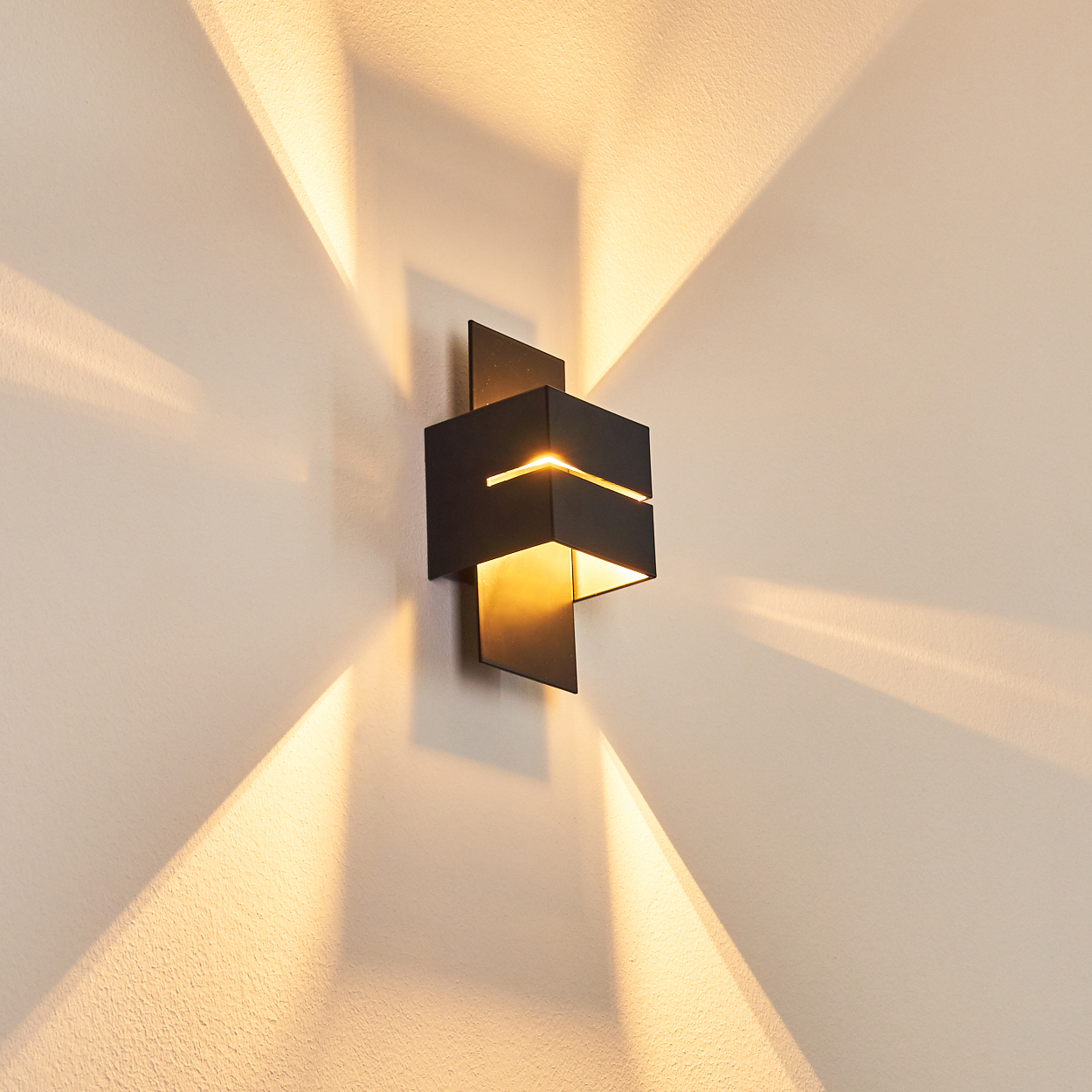 Eine moderne schwarze Wandleuchte mit warmem Licht. Sie wirft ein kreuzförmiges Muster an die beige Wand.