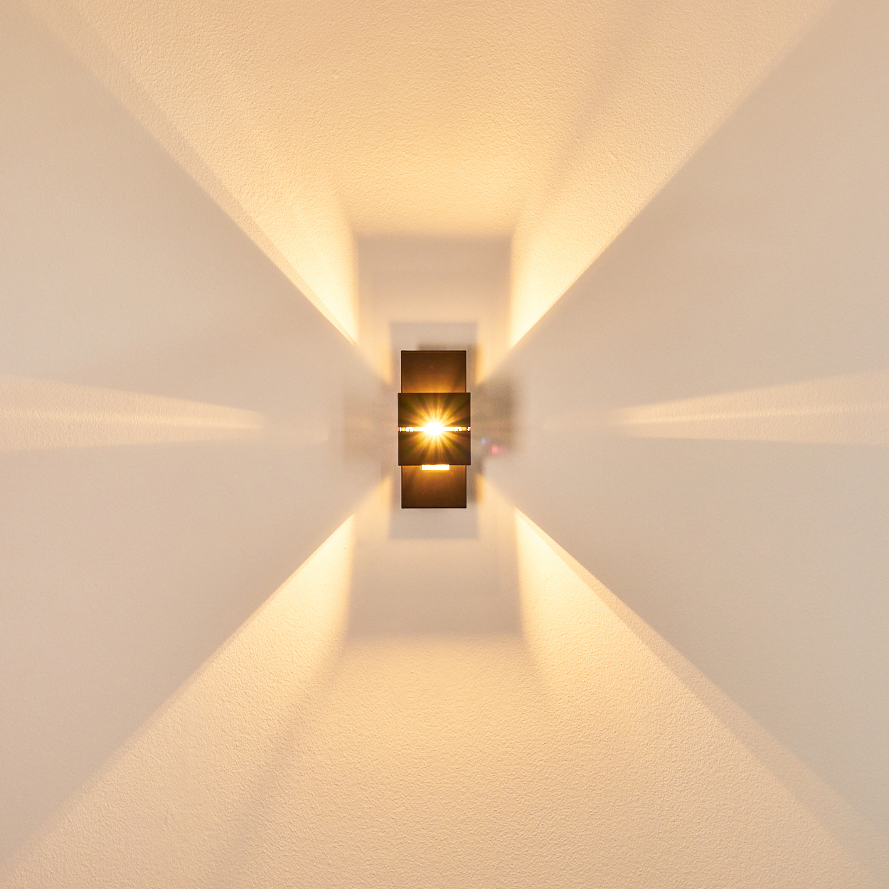 Eine moderne Wandlampe mit drei schwarzen, rechteckigen Teilen. Sie strahlt helles Licht aus und wirft Schatten auf die weiße Wand.