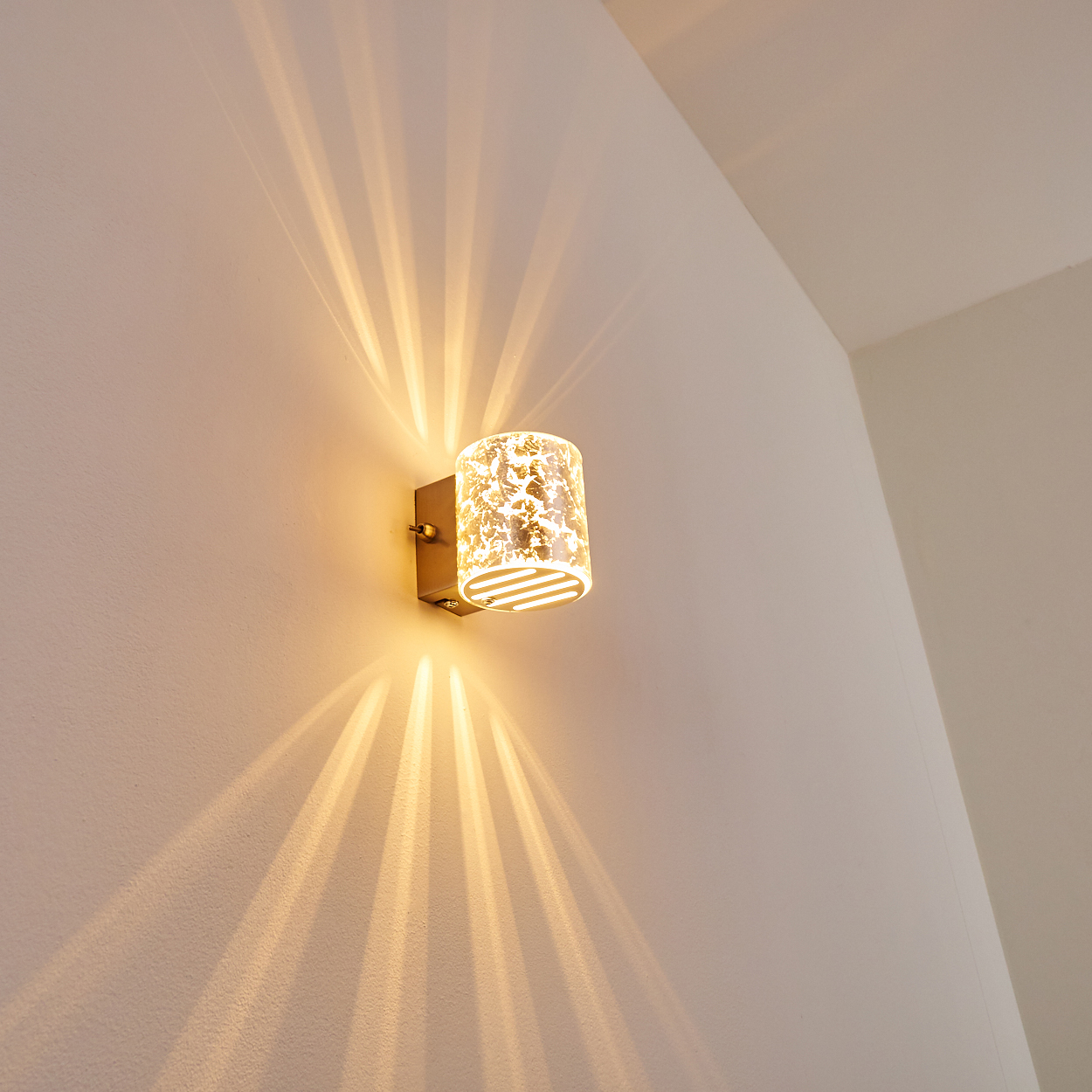 Eine Wandleuchte mit zylindrischem, gemustertem Schirm strahlt Licht aus. Das Licht erzeugt strahlende Muster an der weißen Wand.