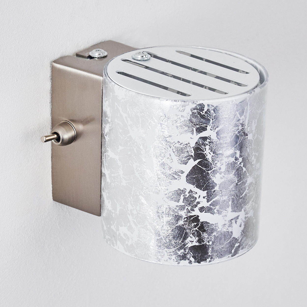 Eine metallische Wandleuchte mit silbernem, gemustertem Schirm, Schalter und Montageplatte an weißer Wand.