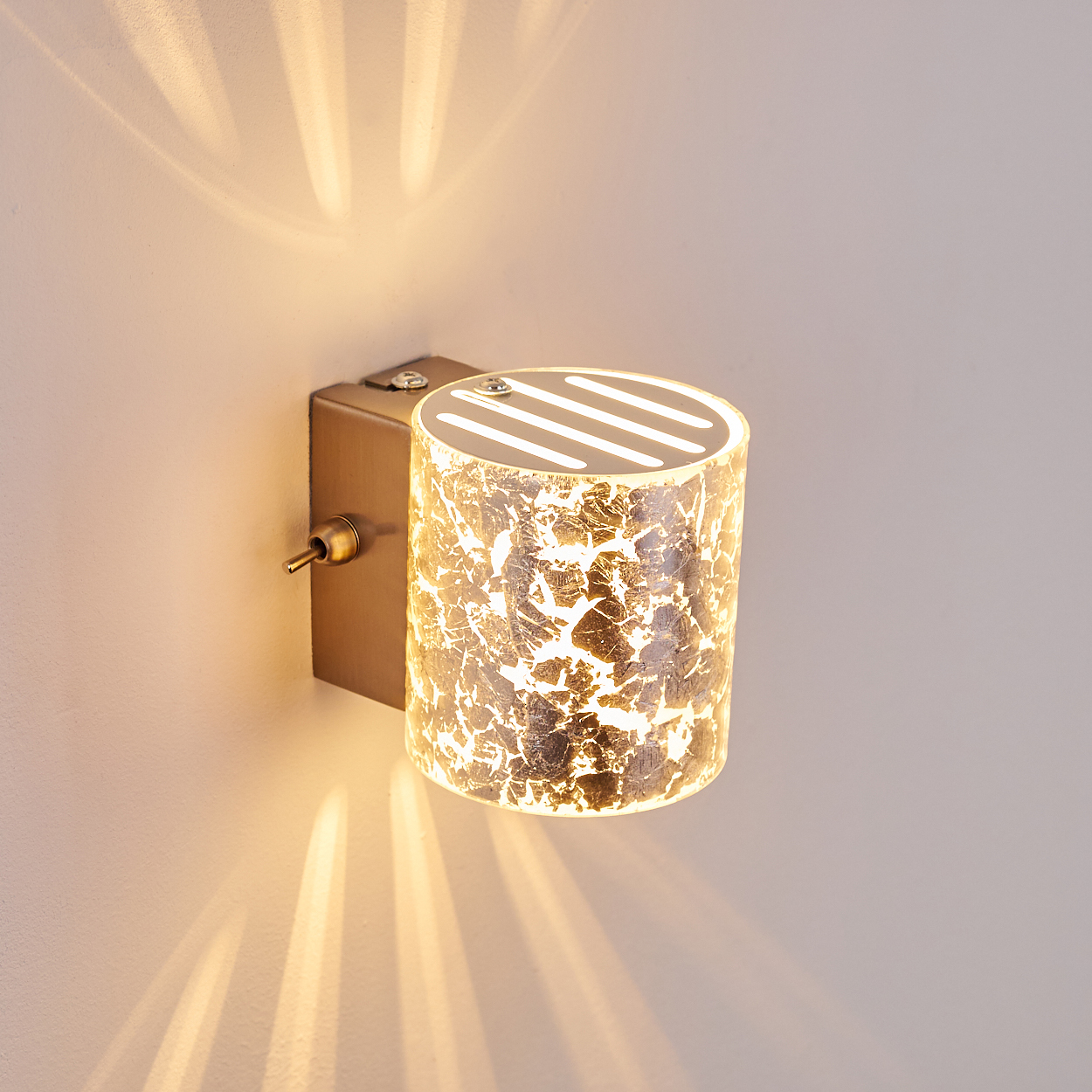 Eine wandmontierte zylindrische Lampe mit strukturiertem Lampenschirm. Das Licht scheint auf die Wand und beleuchtet sie. Die Lampe ist goldfarben.