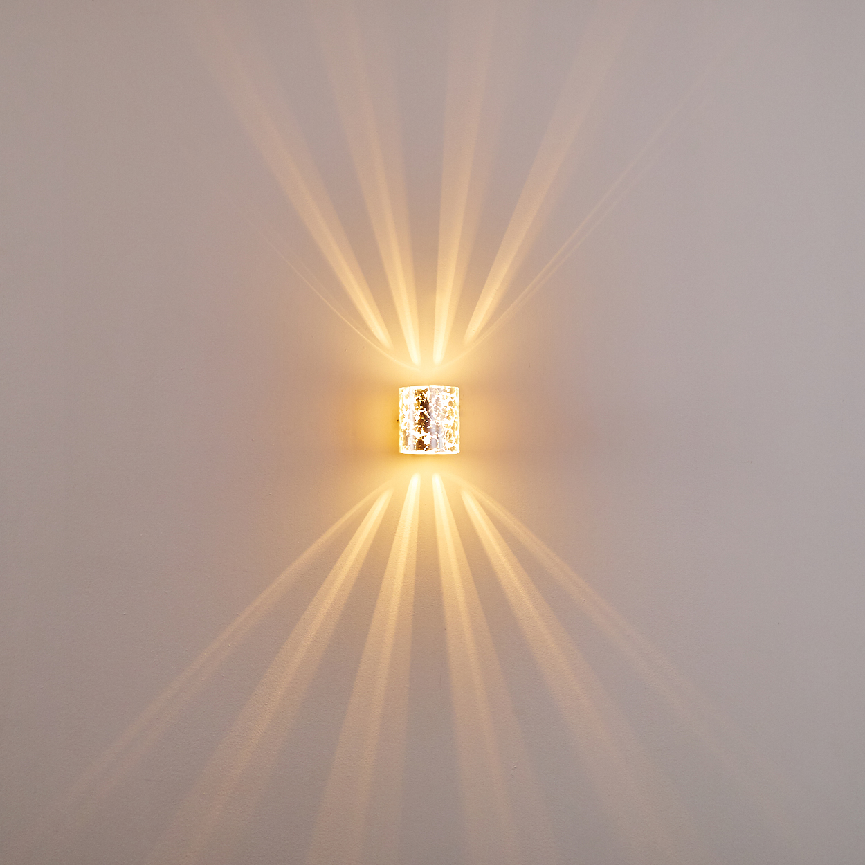 Eine dekorative Wandleuchte strahlt ein warmes Licht aus und wirft radiale Schatten auf eine hellgraue Wand. Die Leuchte ist rechteckig.