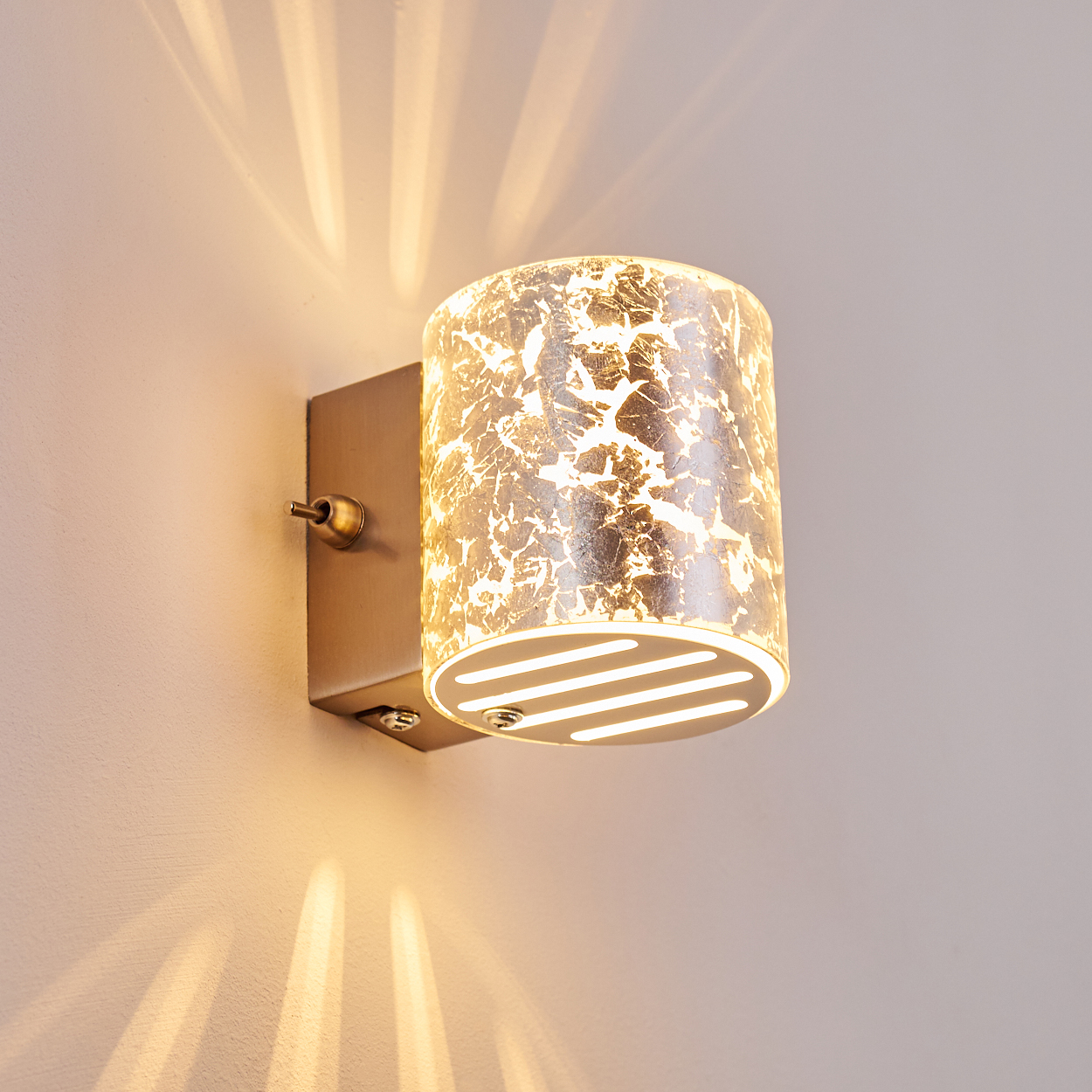Eine Wandlampe mit zylindrischem Lampenschirm. Der Lampenschirm hat ein goldenes, strukturiertes Muster. Die Lampe befindet sich an einer Wand mit Lichtreflexionen.