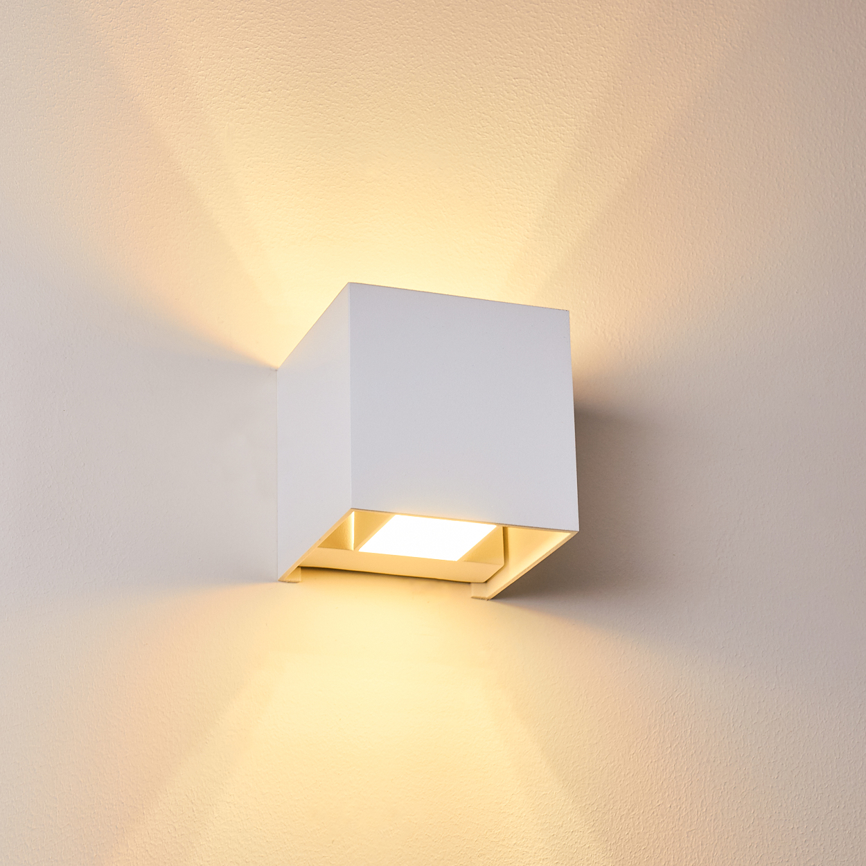 Eine weiße, würfelförmige Wandleuchte beleuchtet eine beige Wand. Licht scheint von der Unter- und Oberseite der Lampe.