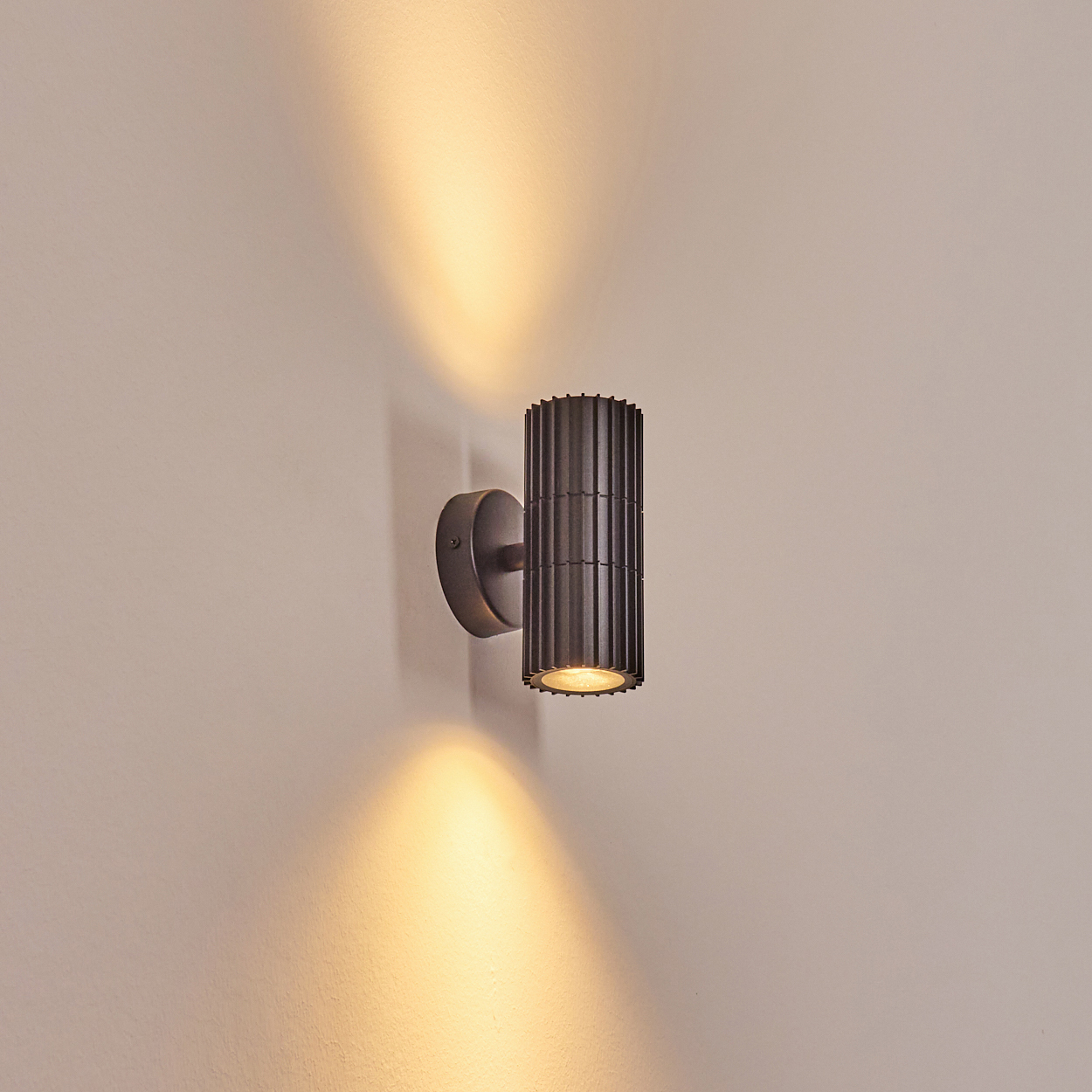 Eine dunkelgraue, zylindrische Wandleuchte mit gerilltem Design ist an einer hellbeige Wand montiert und wirft warmes Licht.
