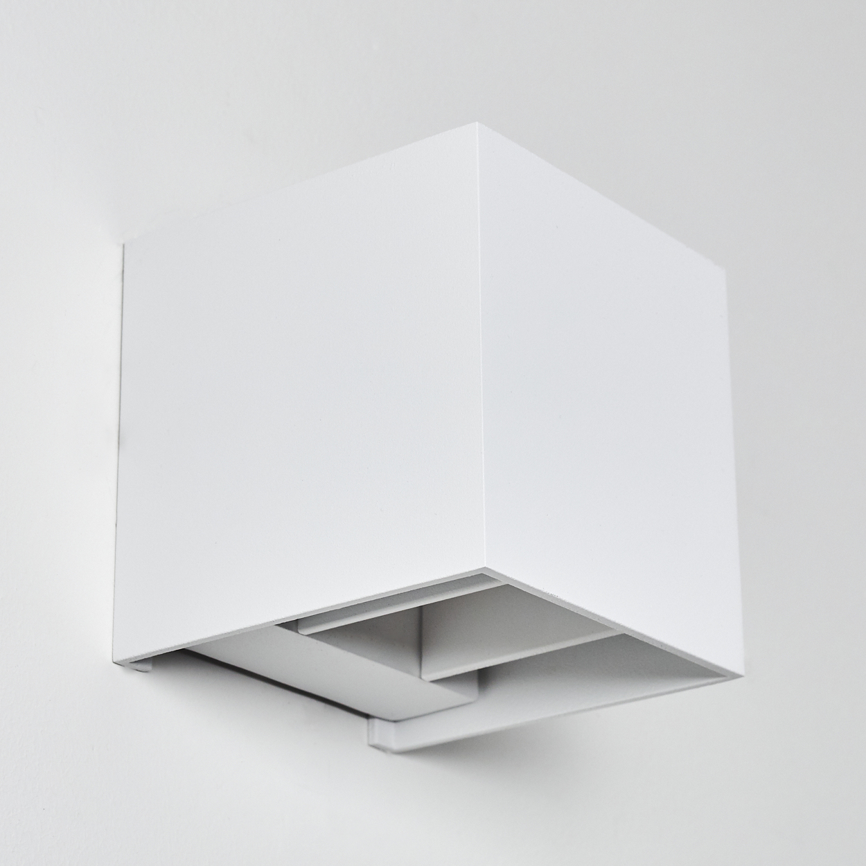 Eine weiße, quaderförmige Wandleuchte ist an einer weißen Wand montiert. Die Lampe hat ein geometrisches Design mit einer vertieften Lichtquelle.