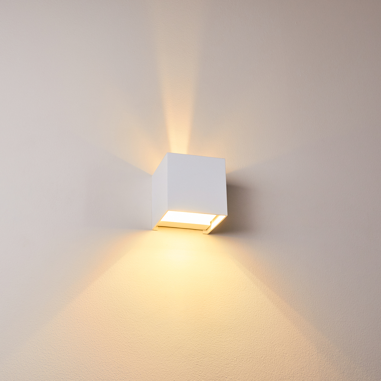 Eine weiße, würfelförmige Wandleuchte beleuchtet eine beige Wand. Licht scheint aus der Lampe und erzeugt Schatten.