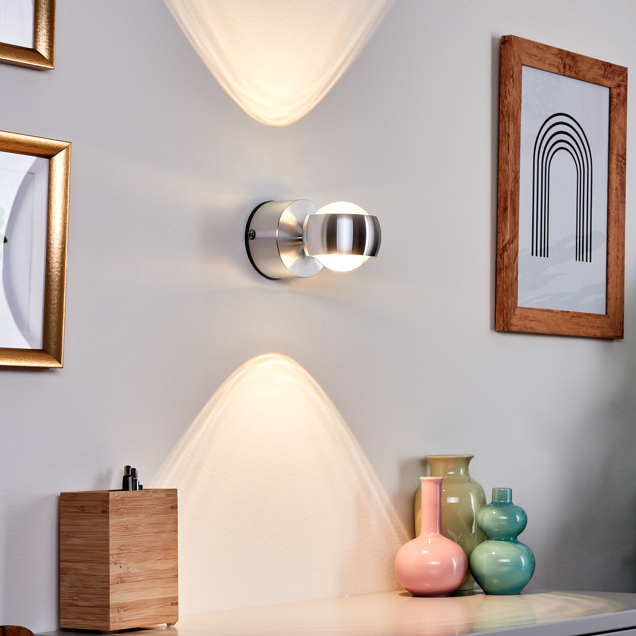 Eine moderne Wandlampe an einer weißen Wand. Darunter Vasen und eine Holzkiste. Gerahmte Bilder sind an der Wand.