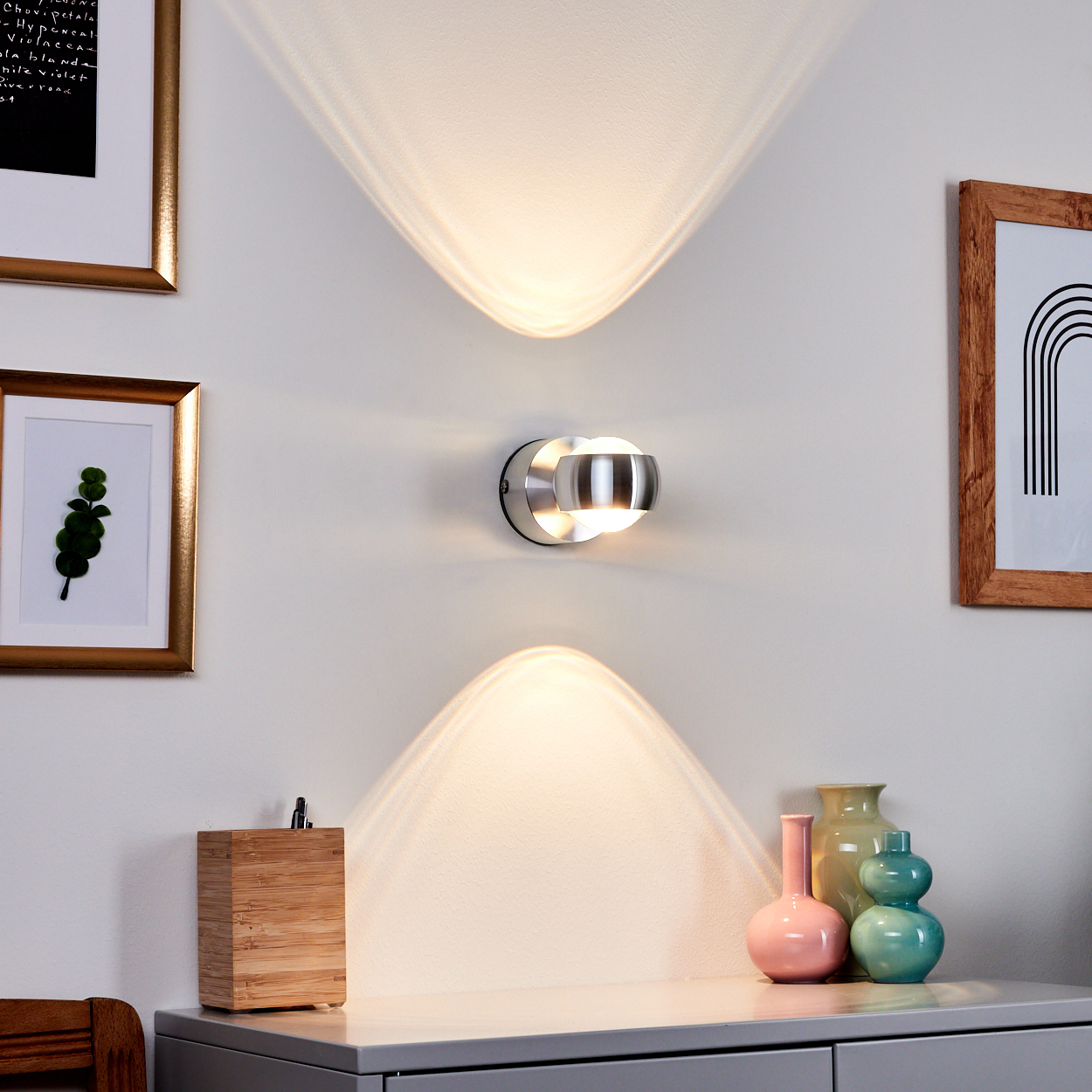Eine moderne Wandlampe wirft Licht auf die Wand. Darunter schmücken eine Holzkiste, Vasen und gerahmte Bilder einen grauen Schrank.