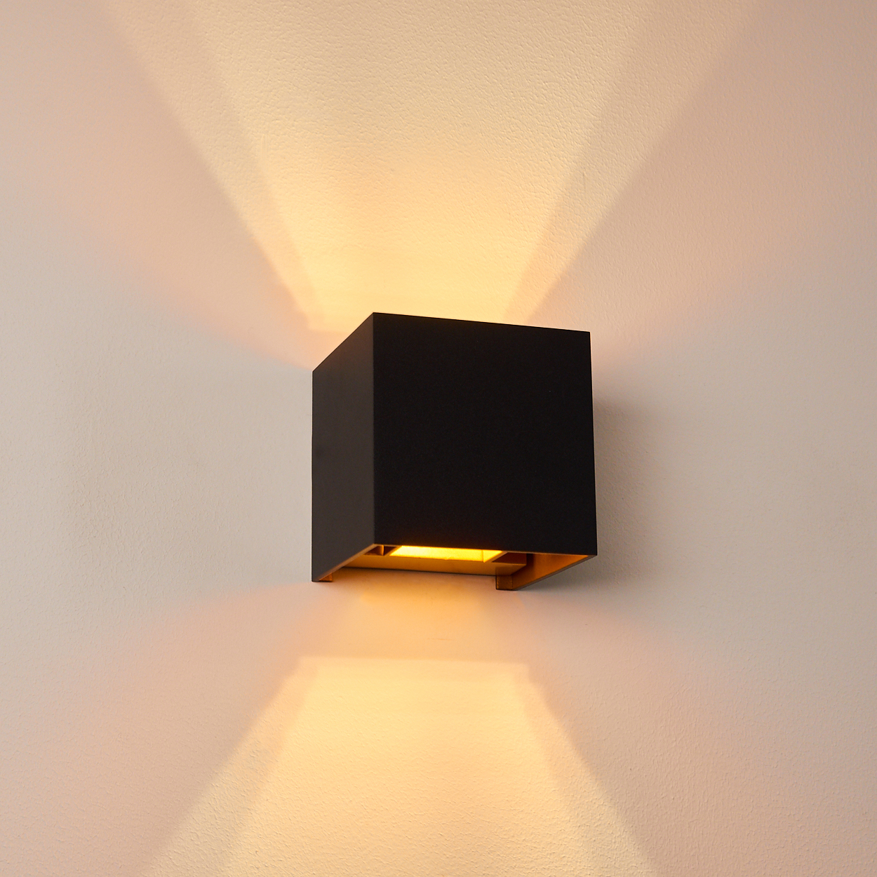 Eine quadratische, schwarze Wandleuchte mit goldenem Innenraum ist an einer weißen Wand montiert. Die Lampe wirft Licht nach oben und unten.