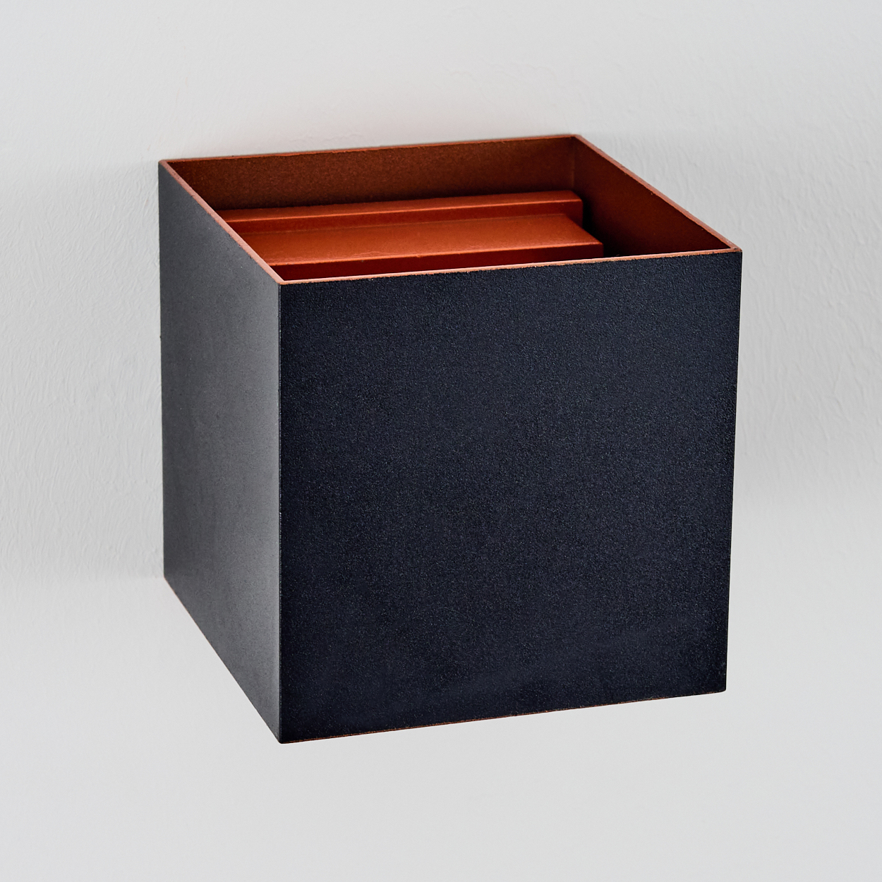 Eine dunkelblaue quadratische Box an einer weißen Wand. Innen und Oberkante sind orange. Die Box ist leicht geneigt.