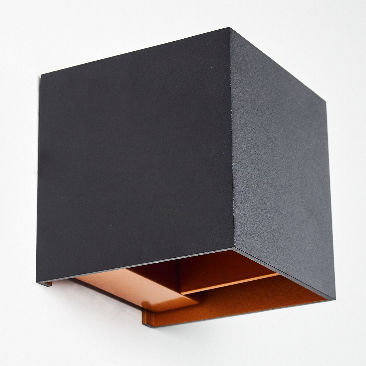 Eine moderne schwarze Würfellampe an einer weißen Wand. Die Innenseite der Lampe ist kupferfarben. Klares und minimalistisches Design.