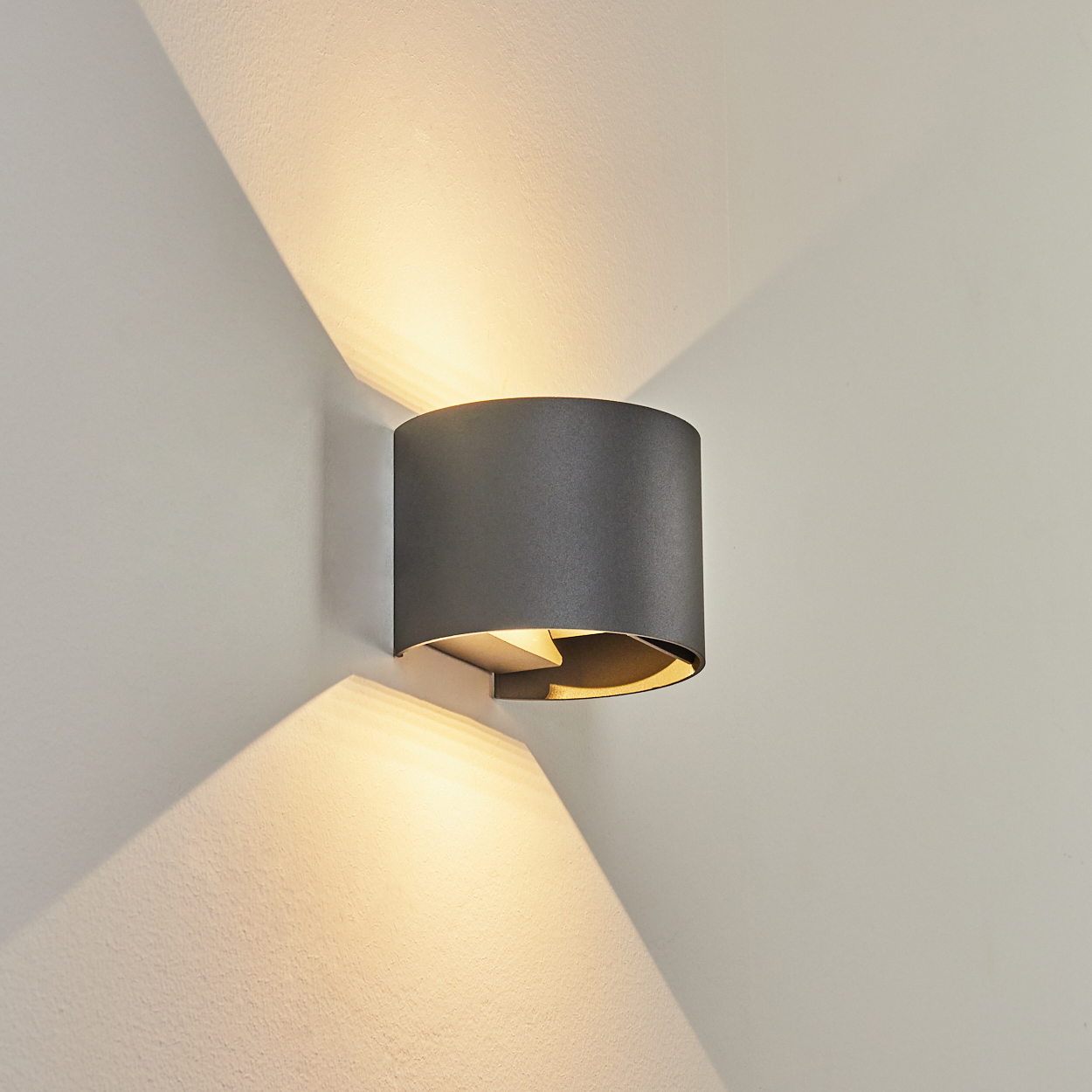 Eine graue zylindrische Wandleuchte ist an einer weißen Wand befestigt. Licht scheint nach oben und unten und erzeugt einen Kontrast von Licht und Schatten.
