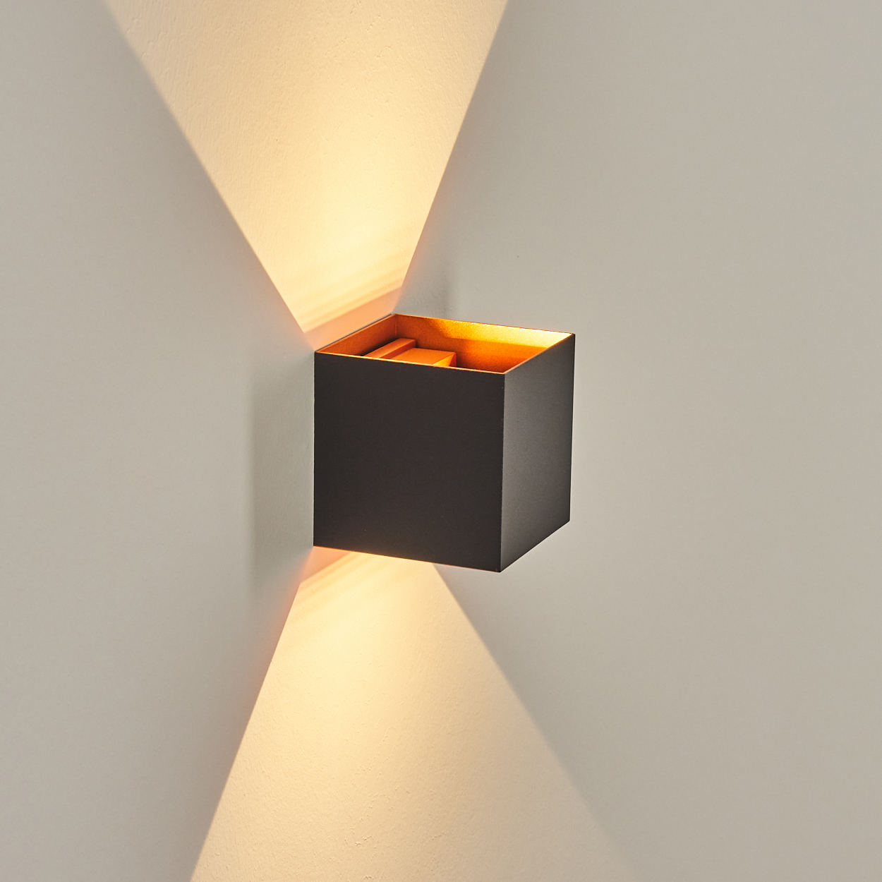 Eine quadratische schwarze Wandleuchte mit goldenem Innenraum. Lichtstrahlen nach außen. An einer weißen Wand montiert. Schatten und Licht sind sichtbar.