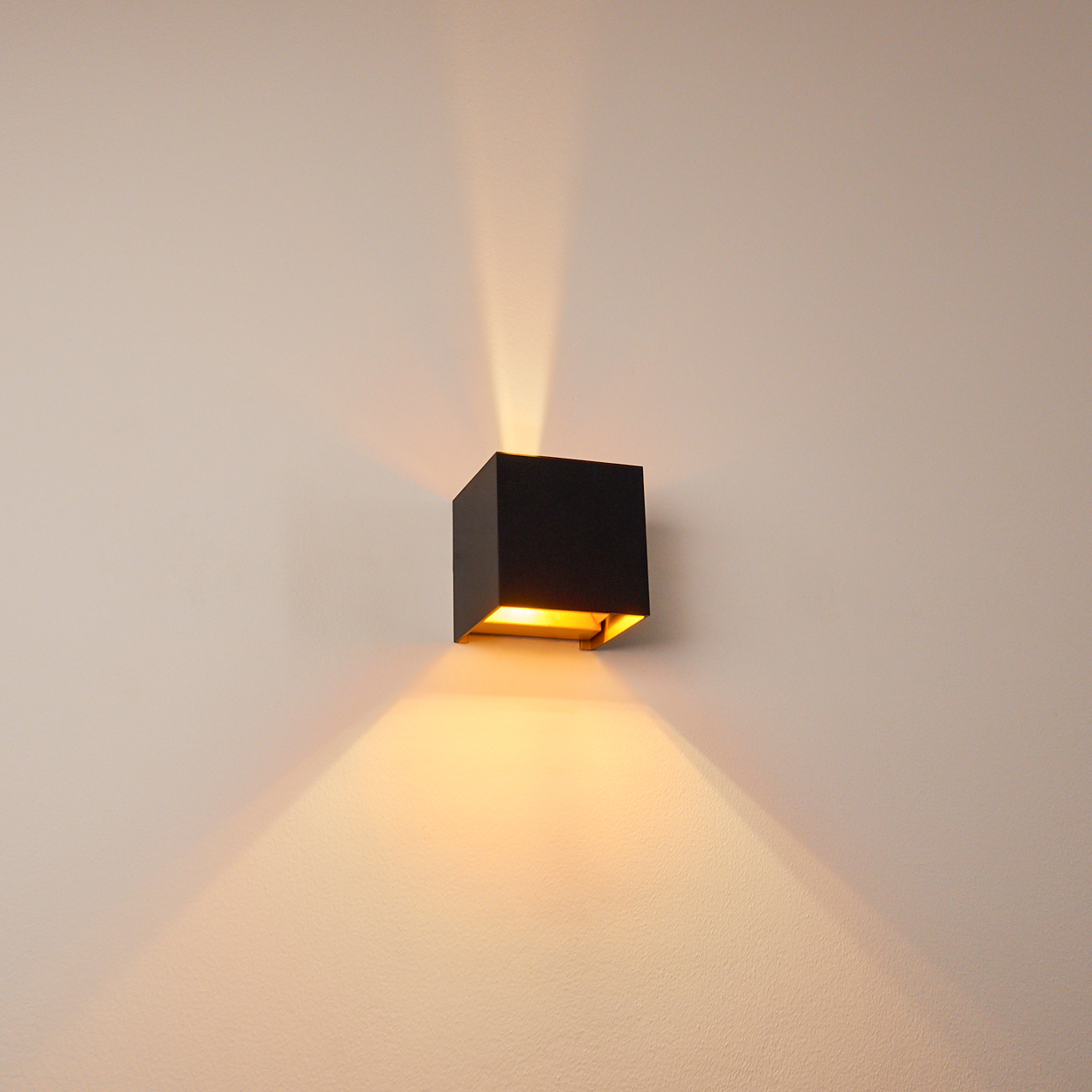 Eine quadratische, schwarze Wandleuchte an einer hellbeige Wand. Die Lampe strahlt helles Licht nach oben und unten aus.