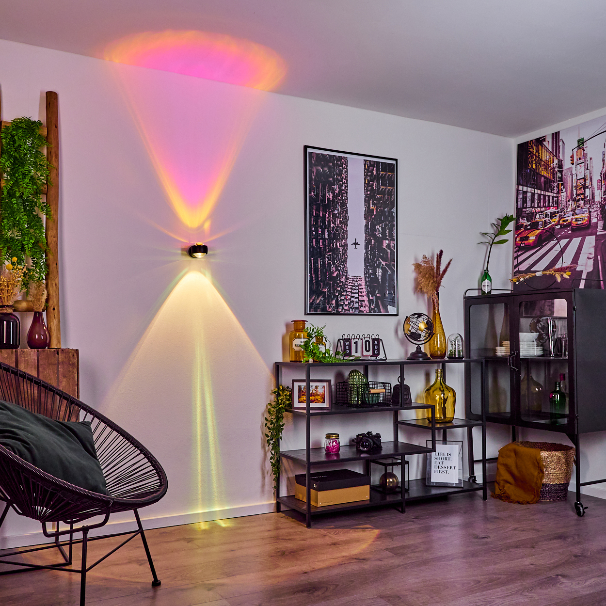 Ein Raum mit einer Wandleuchte, die farbenfrohes Licht projiziert. Es gibt einen schwarzen Stuhl, Regale und gerahmte Bilder. Ein NYC-Kunstwerk ist sichtbar.