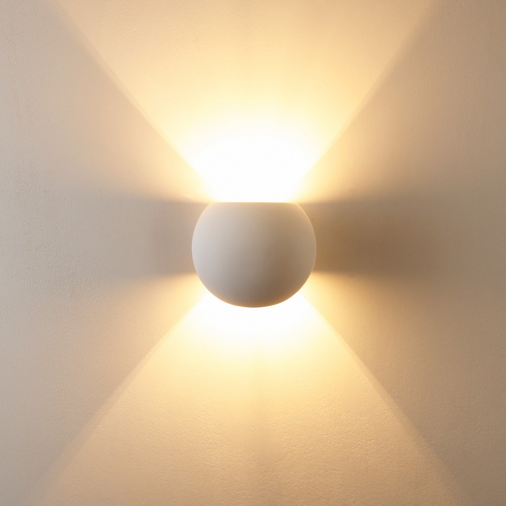 Eine weiße, kugelförmige Wandleuchte an einer strukturierten, hellfarbigen Wand. Die Lampe strahlt helles Licht nach oben und unten aus.