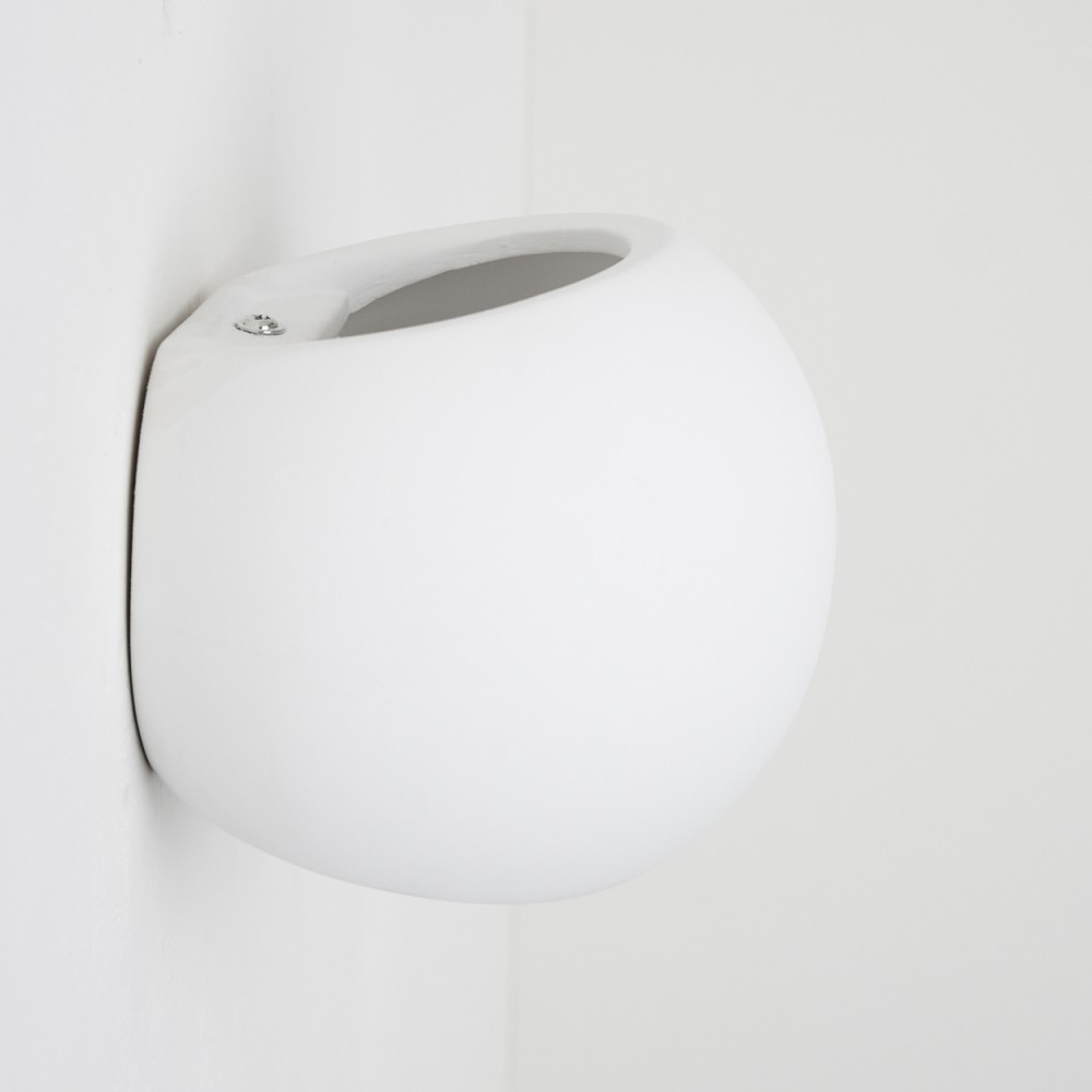 Ein weißer, kugelförmiger Wandtopf. Er befindet sich in einer Ecke und ist mit einer Schraube befestigt. Die Öffnung ist oval geformt.