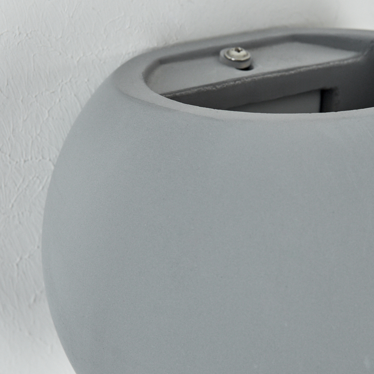 Nahaufnahme einer grauen, abgerundeten Wandleuchte. Sie befindet sich vor einer strukturierten weißen Wand, mit sichtbarem Schraubendetail.