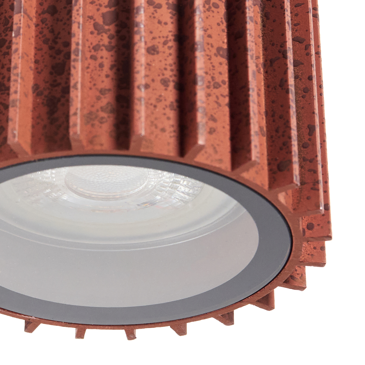 Nahaufnahme einer rostfarbenen zylindrischen Leuchte. Sie hat ein eingelassenes Licht und ein strukturiertes Metallgehäuse mit weißem Hintergrund.