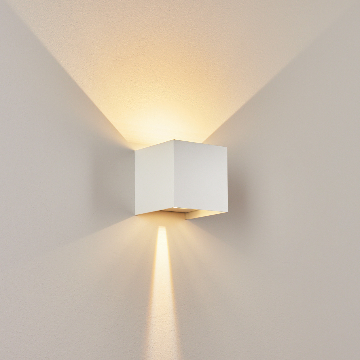 Eine weiße quadratische Wandleuchte strahlt Licht nach oben, unten und zu den Seiten und wirft Schatten auf die beige Wand.
