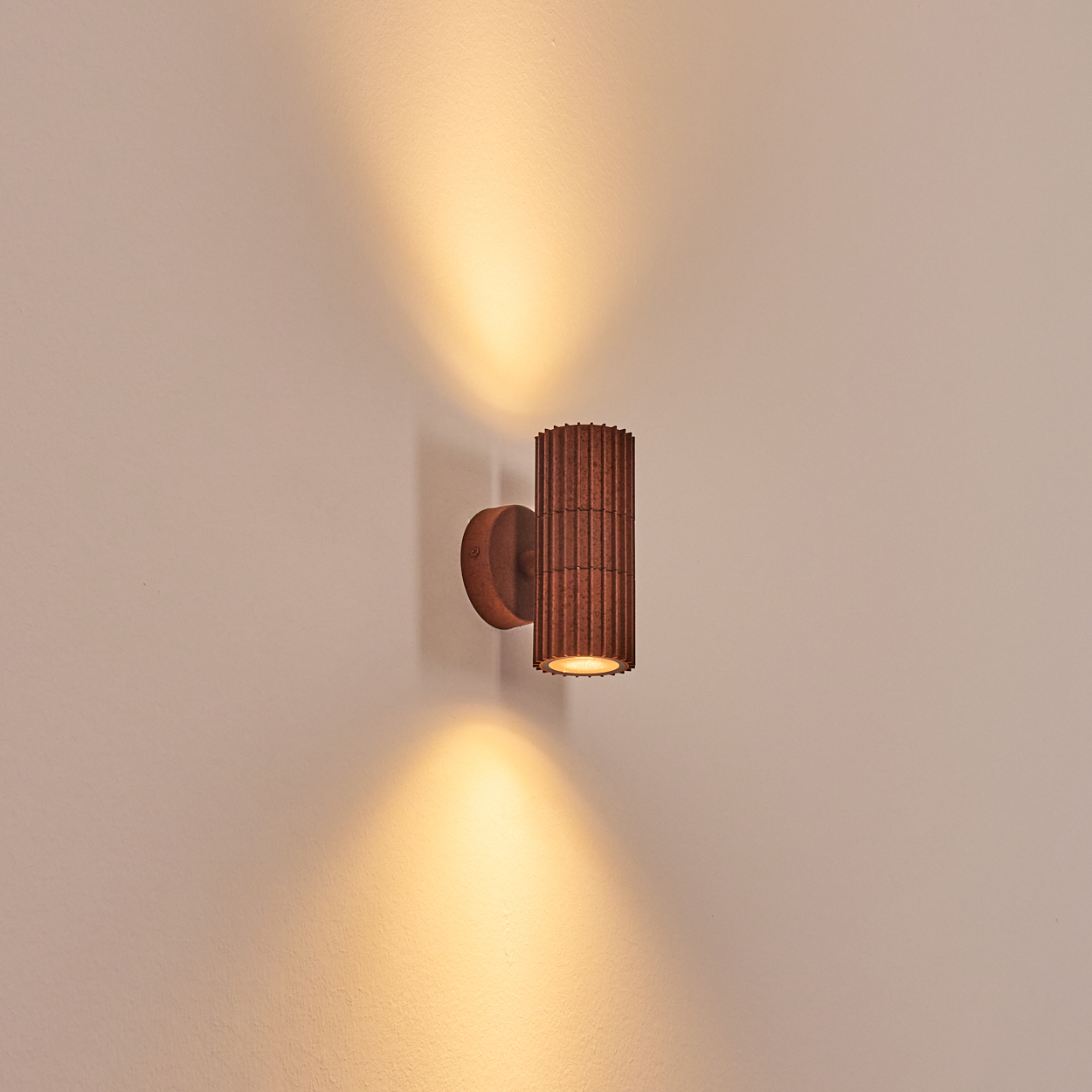 Eine braune Wandleuchte ist an einer hellen Wand montiert. Die Lampe strahlt Licht nach oben und unten aus. Die Wand hat eine schlichte Textur.