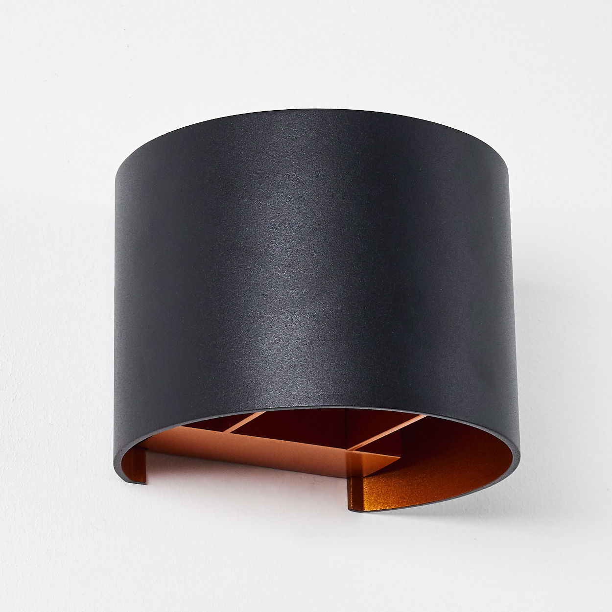 Eine moderne schwarze Wandleuchte mit kupferfarbenem Innenraum ist an einer weißen Wand montiert. Die Lampe hat eine zylindrische Form.