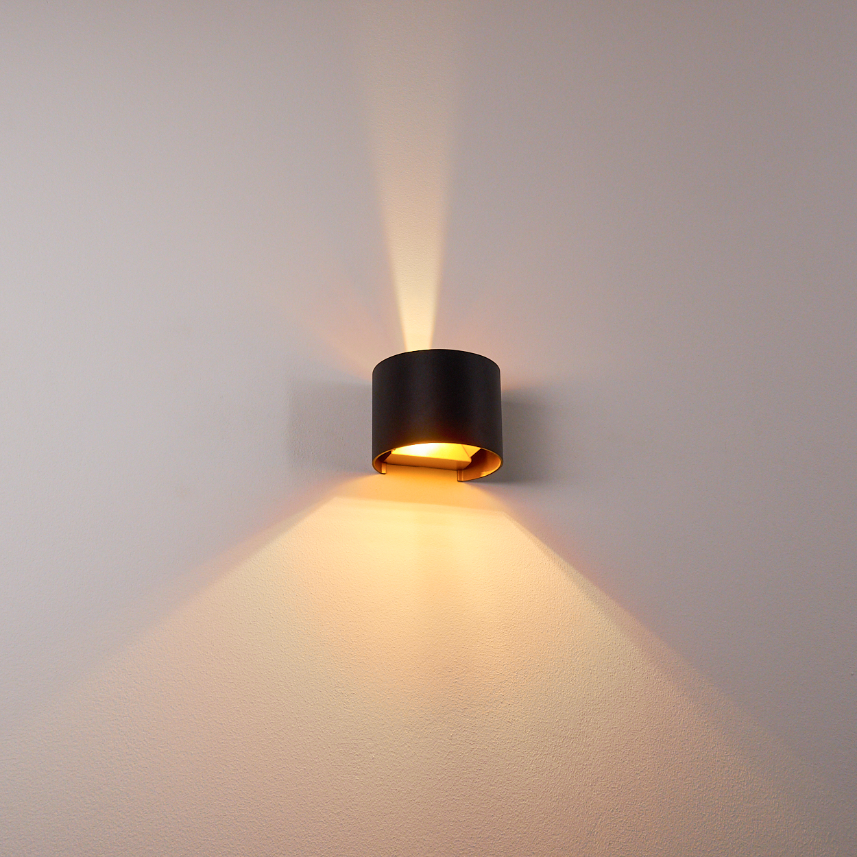 Eine schwarze, zylindrische Wandleuchte strahlt warmes Licht aus. Das Licht beleuchtet die Wand und erzeugt ein Muster aus Licht und Schatten.
