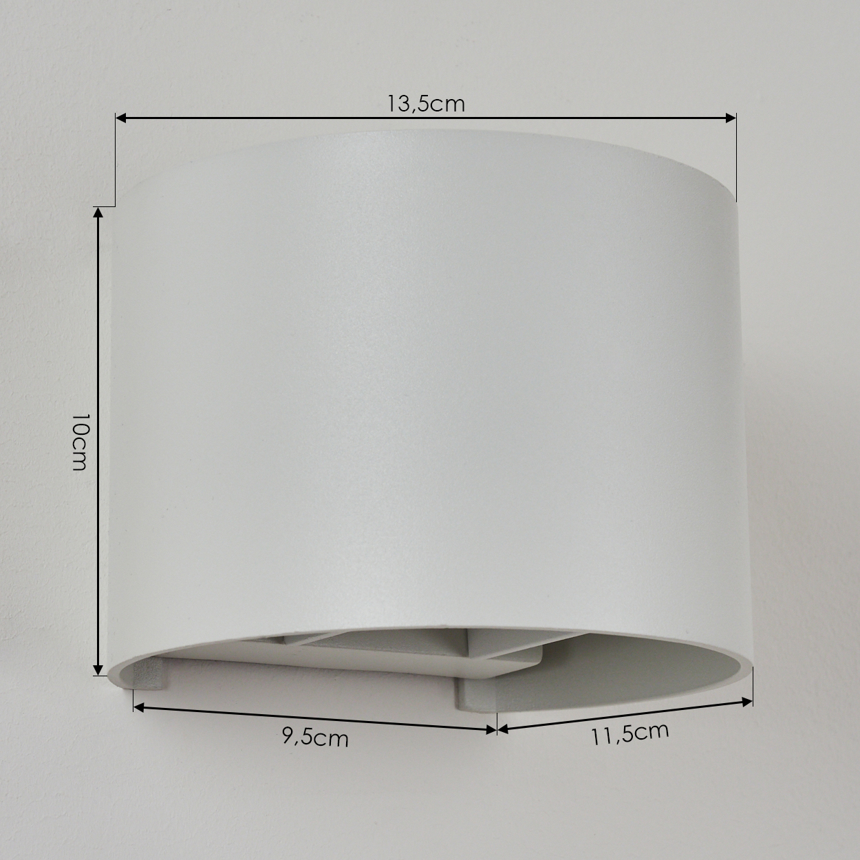 Weiße, zylindrische Wandleuchte. Abmessungen: 13,5 cm breit, 10 cm hoch, Sockel 9,5 cm und 11,5 cm. An einer weißen Wand montiert.