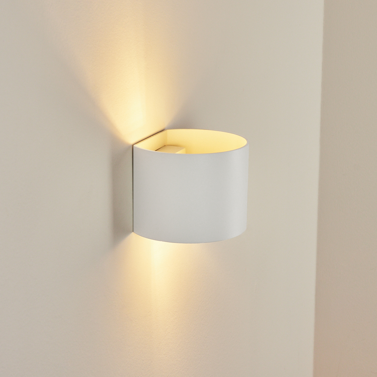 Eine weiße Wandleuchte beleuchtet die Wand. Die Lampe ist zylindrisch mit warmem, gelbem Licht. Die Wand ist beige.