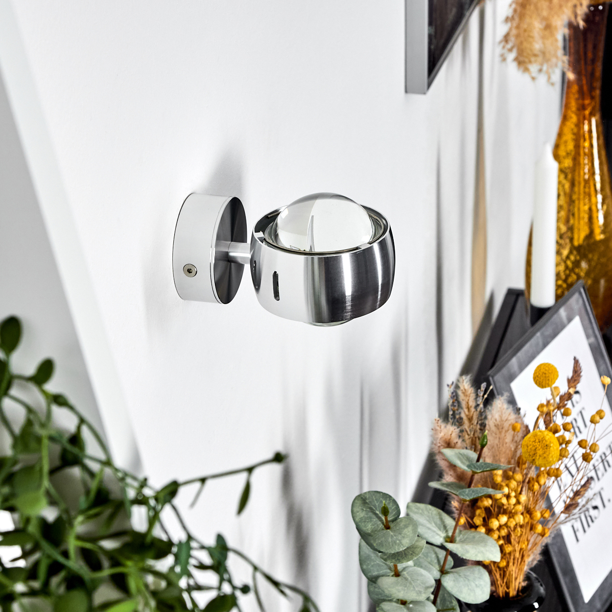 Eine moderne Wandlampe aus Chrom mit Glasabdeckung ist an einer weißen Wand befestigt. Dekorative Gegenstände, darunter Blumen, sind sichtbar.