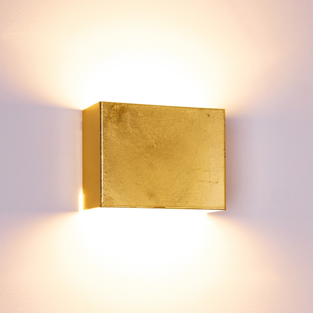 Eine goldene quadratische Wandleuchte ist an einer weißen Wand montiert. Die Lampe strahlt nach oben und unten Licht aus.