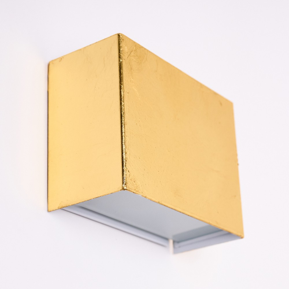 Eine goldene, rechteckige Wandleuchte ist an einer weißen Wand montiert. Die Lampe hat eine kastenförmige Form mit sichtbarer Textur und hellem Innenraum.