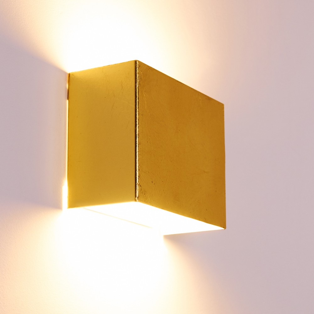 Eine goldene, würfelförmige Wandleuchte beleuchtet die Wand. Licht scheint von unten und den Seiten und wirft Schatten auf eine weiße Wand.