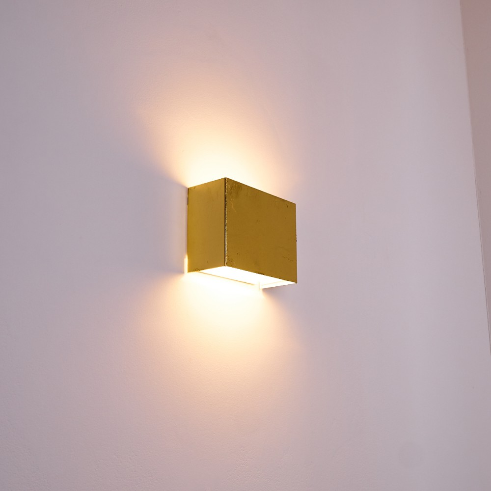 Eine goldene, würfelförmige Wandleuchte beleuchtet eine weiße Wand. Das Licht scheint nach oben und unten und erzeugt Schatten.