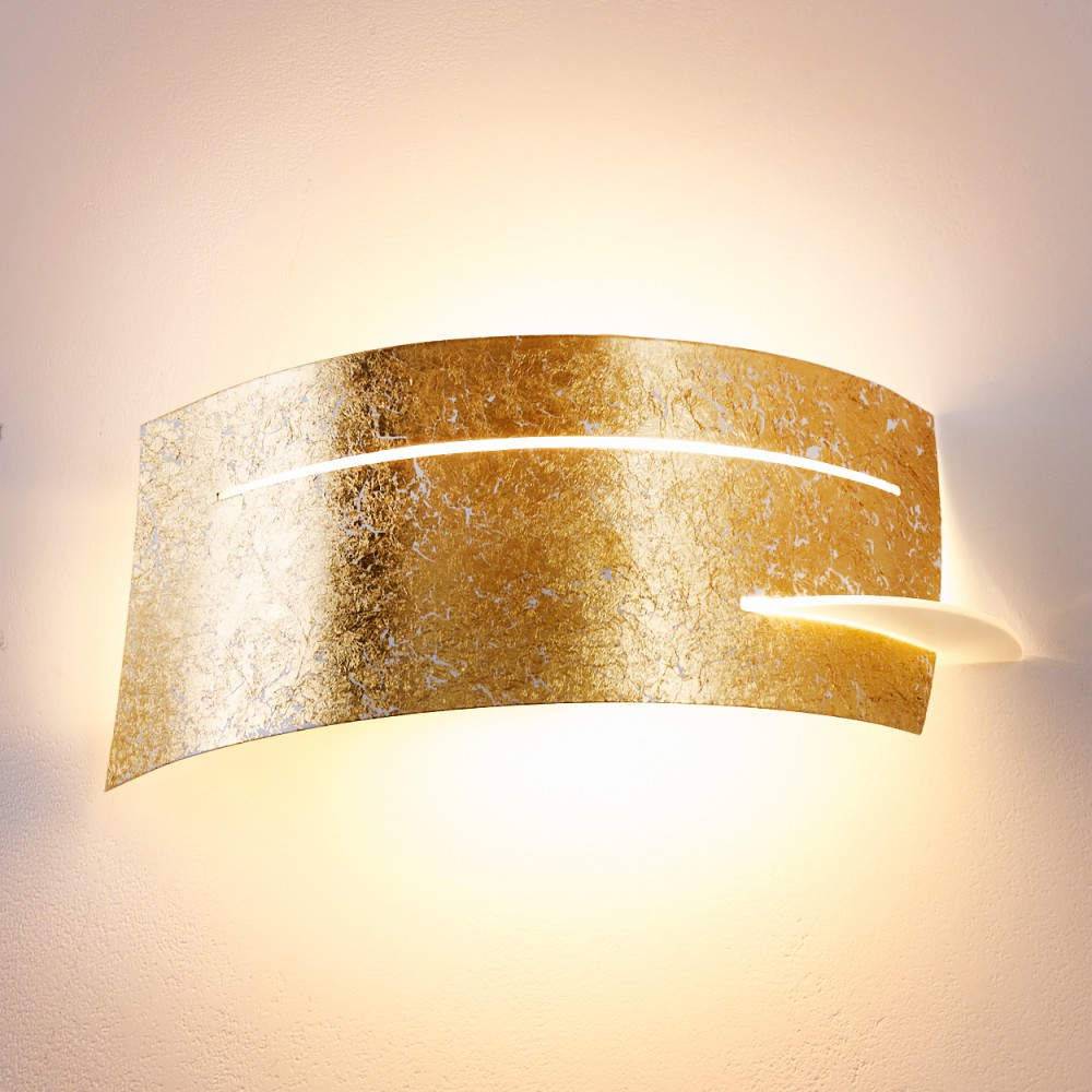 Eine goldene Wandlampe mit geschwungenem Design, strukturiertem Blattgold und warmem, diffusem Licht an einer weißen Wand.