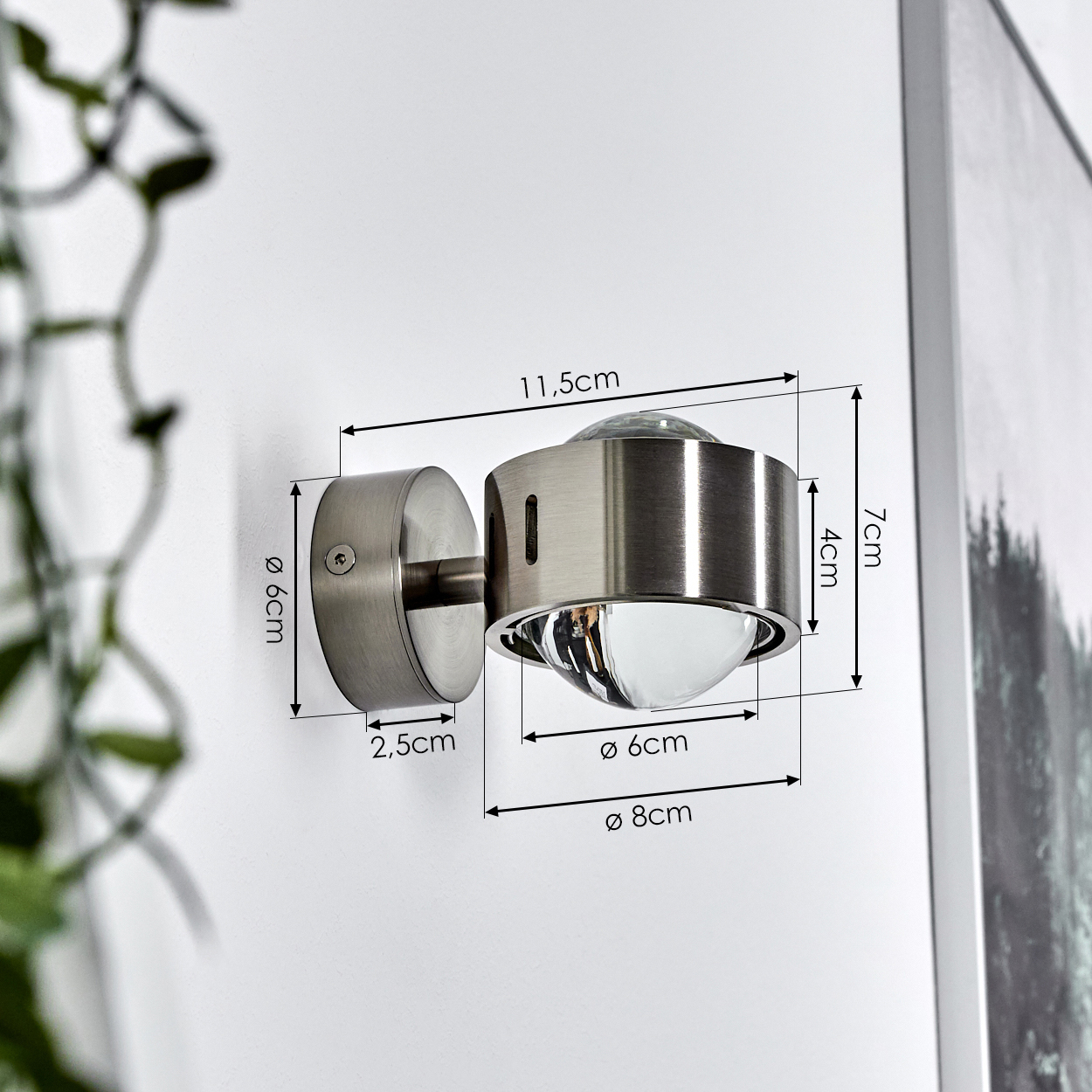 Eine metallische Wandleuchte ist an einer weißen Wand befestigt. Abmessungen sind auf der Lampe angegeben. Rechts ist ein Gemälde zu sehen.