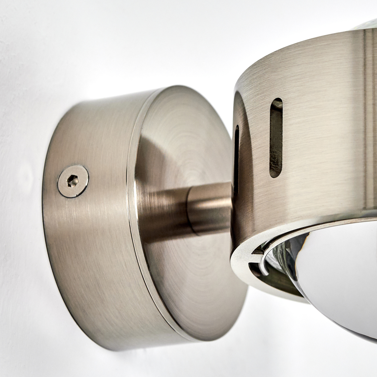 Nahaufnahme einer Wandlampe aus gebürstetem Metall. Die Lampe hat einen runden Sockel, einen zylindrischen Arm und ein Glühbirnengehäuse.