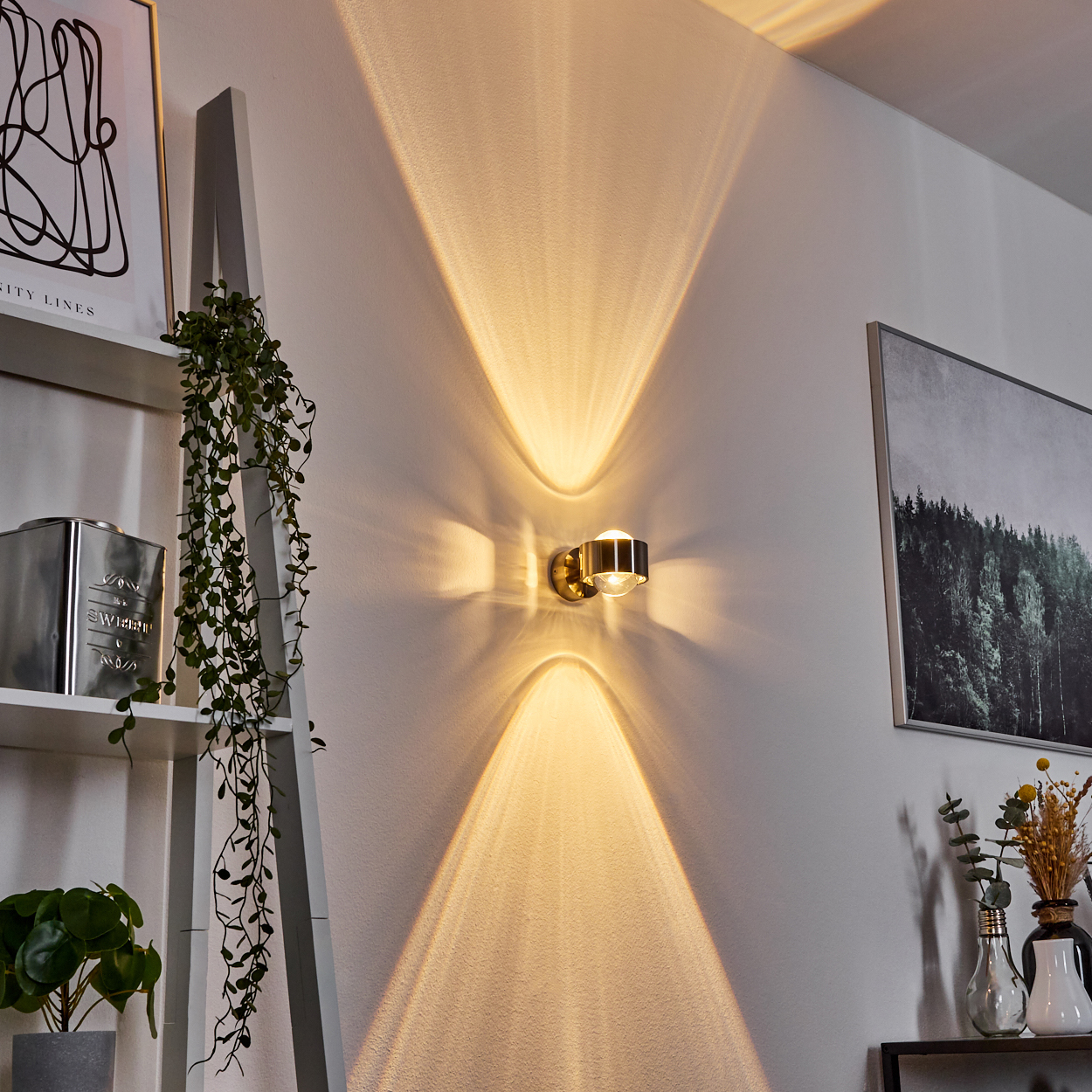Eine goldene Wandlampe wirft Licht nach oben und unten. Auf der linken Seite befinden sich ein Leiterregal mit Pflanzen und ein Bilderrahmen.