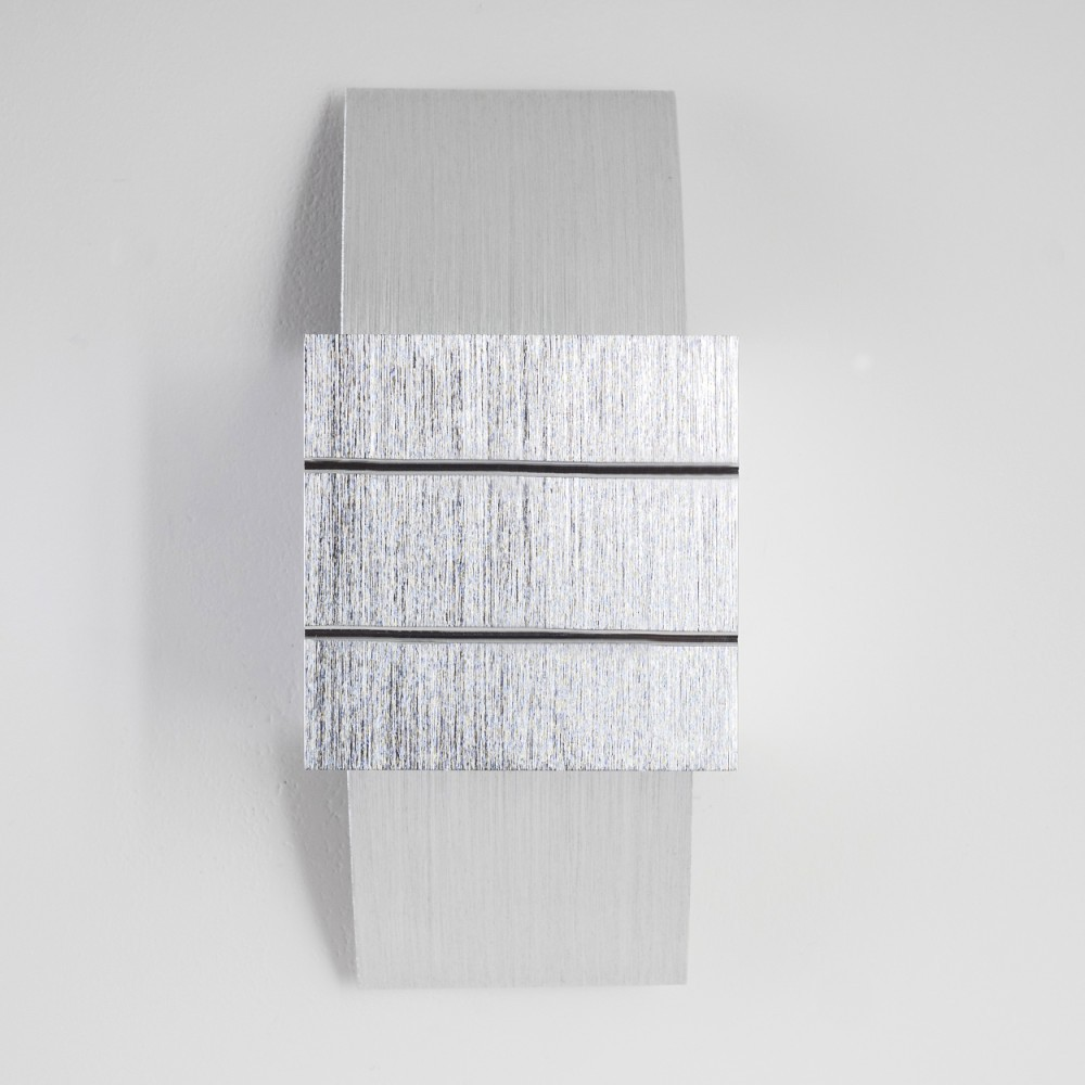 Eine metallische Wanddekoration mit einem quadratischen Mittelteil, strukturiert mit vertikalen Linien, und einem glatten Rechteck.